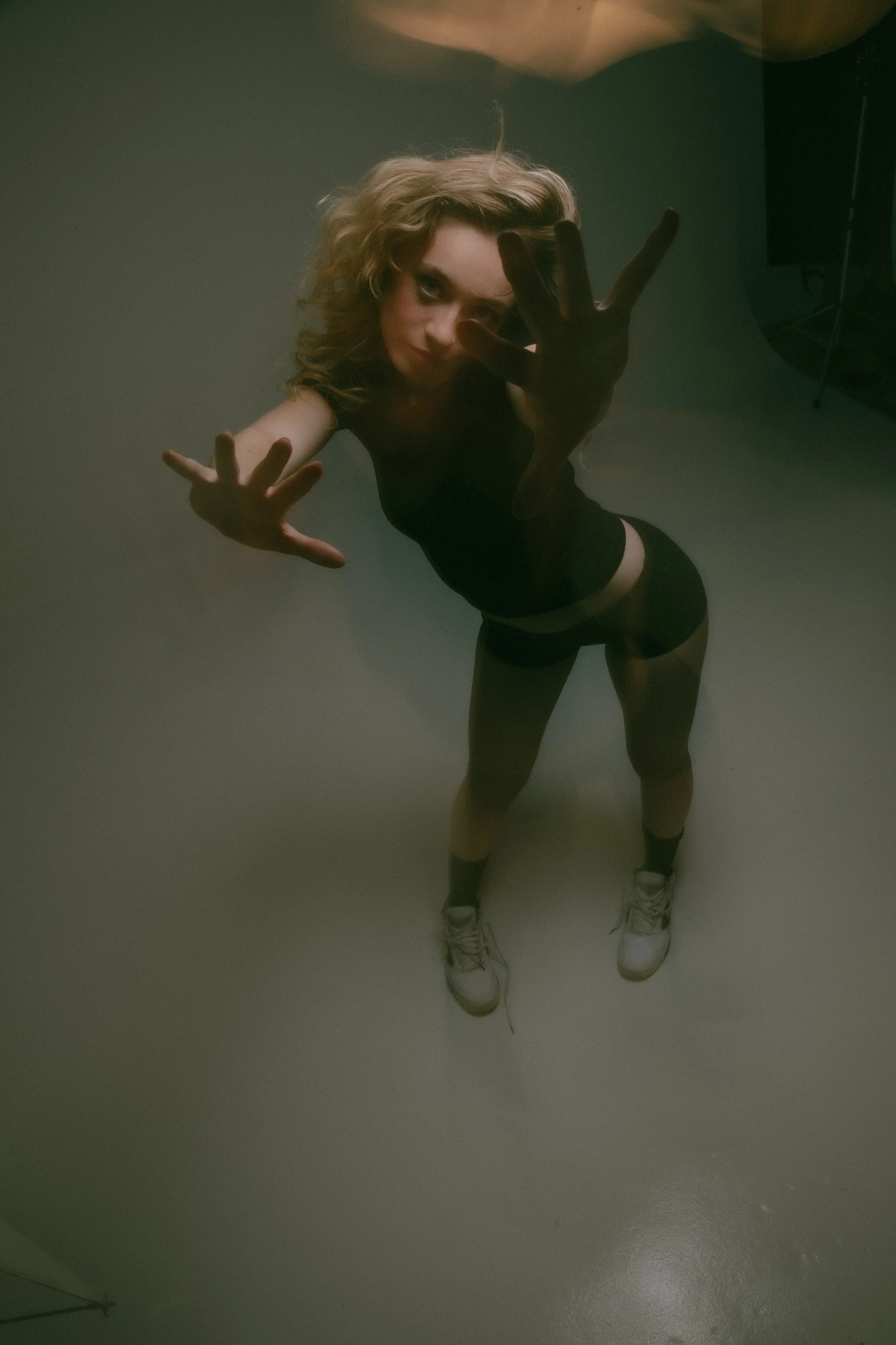 A young woman with curly hair taking a photo of herself through a reflective surface, reaching out toward the camera with one hand while standing on a plain floor.