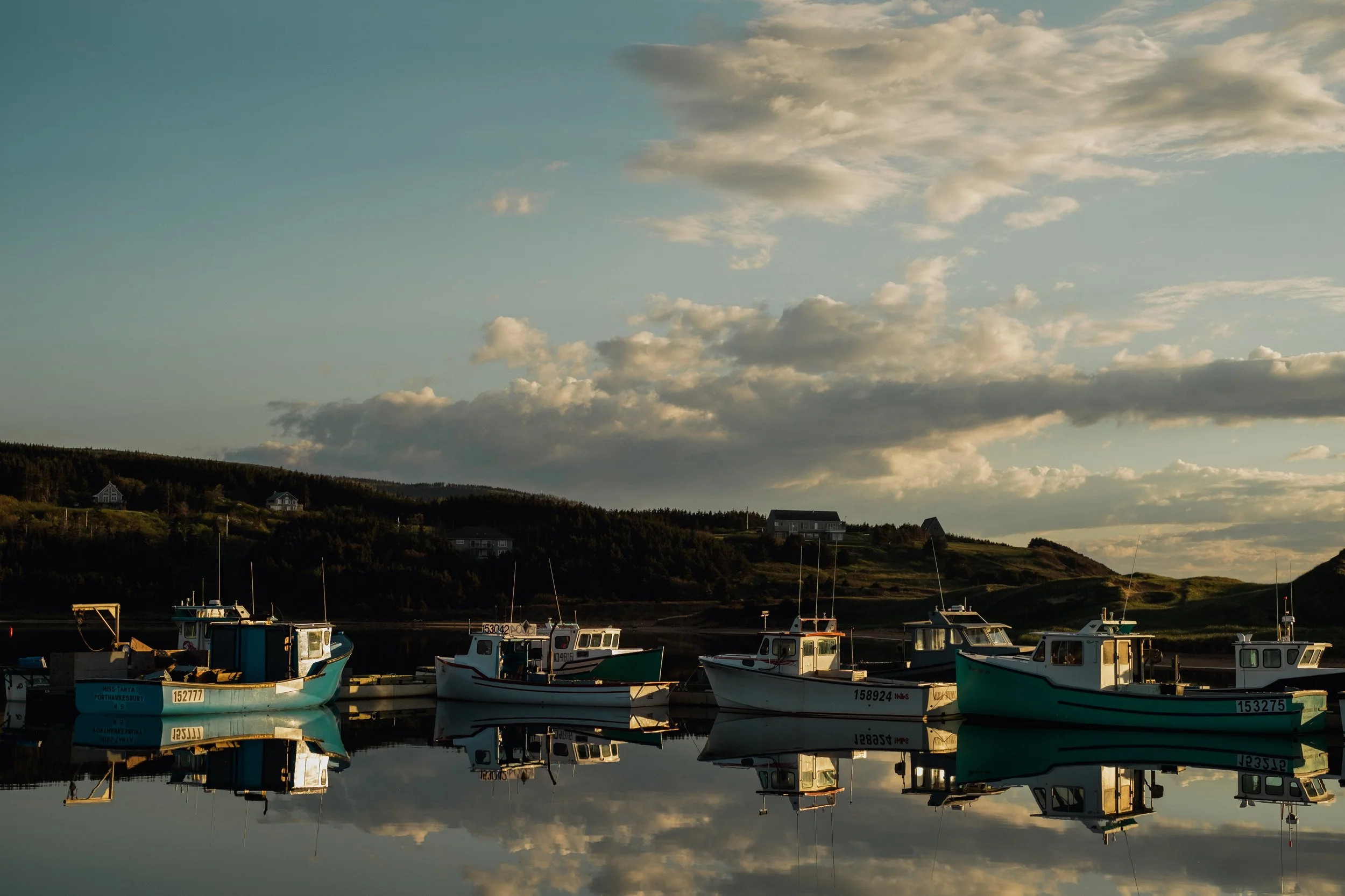 Several boats are docked in calm water with reflections, hills with houses in the background, and a partly cloudy sky during sunset.