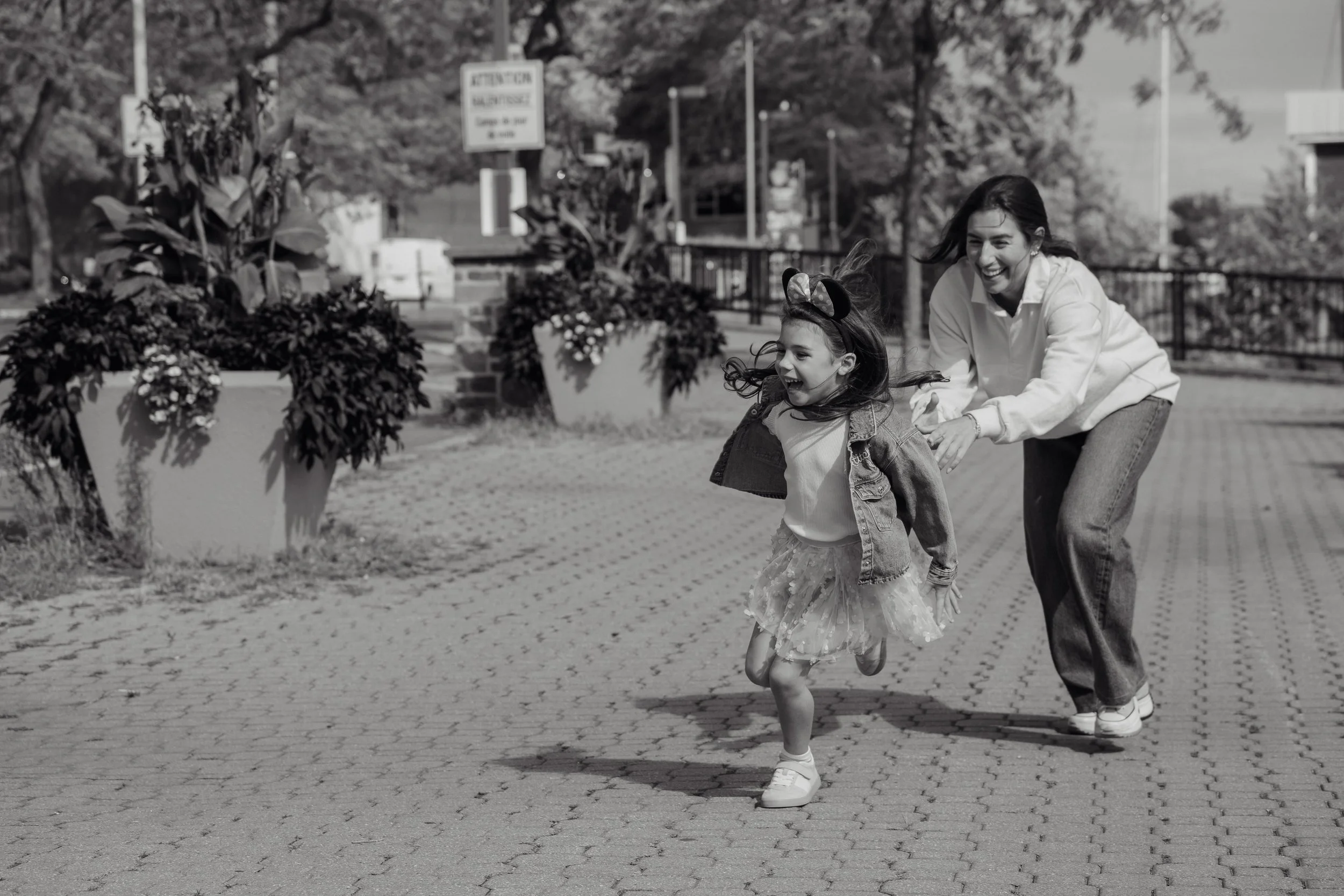 A woman and a young girl are playing and running outdoors on a paved area, ahead of a garden with potted plants, in a park or city area, with trees and buildings in the background. The woman is wearing a white sweatshirt and jeans, and the girl is we