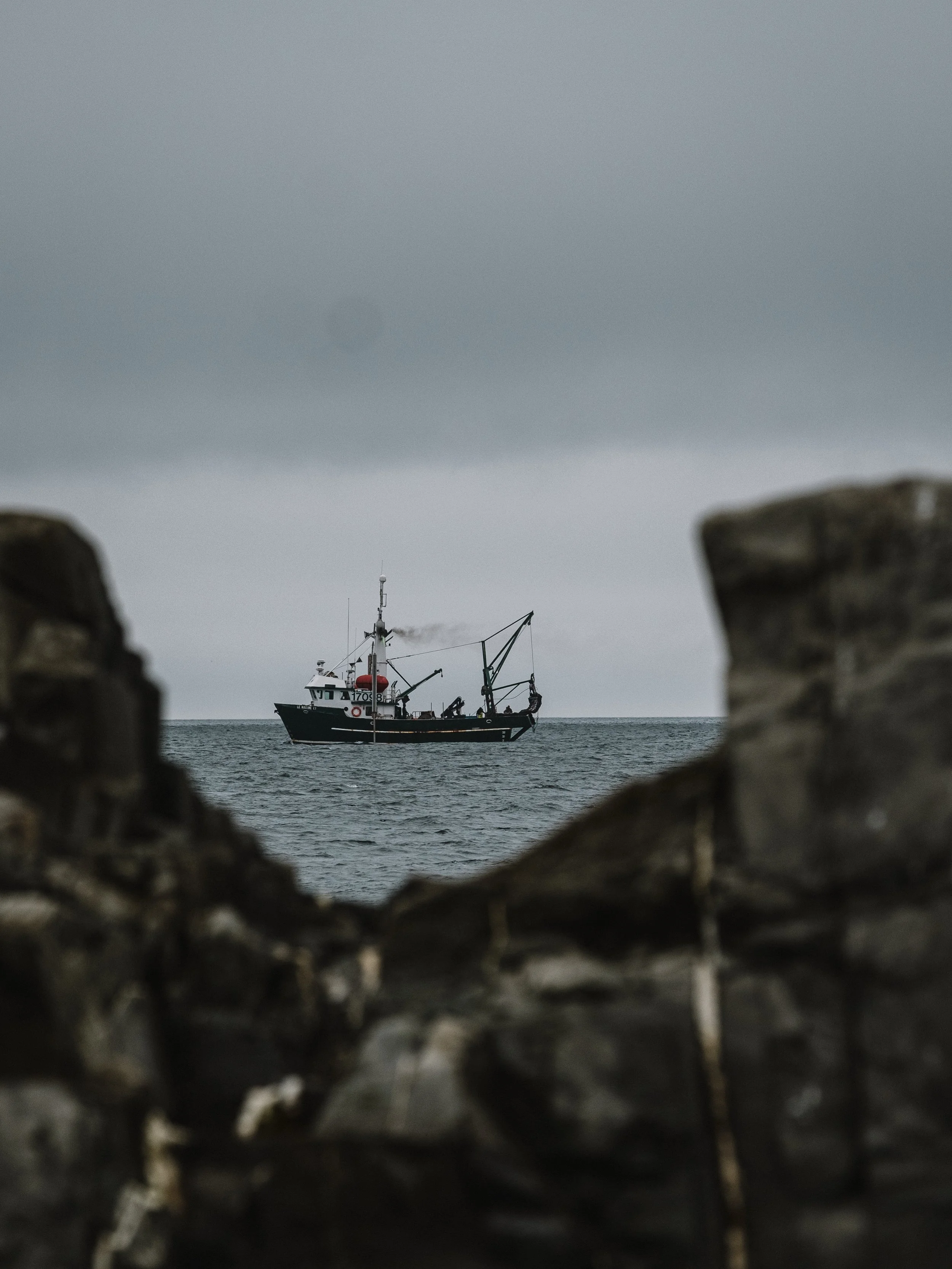 A fishing boat on the ocean seen through a gap in rocks during an overcast day.