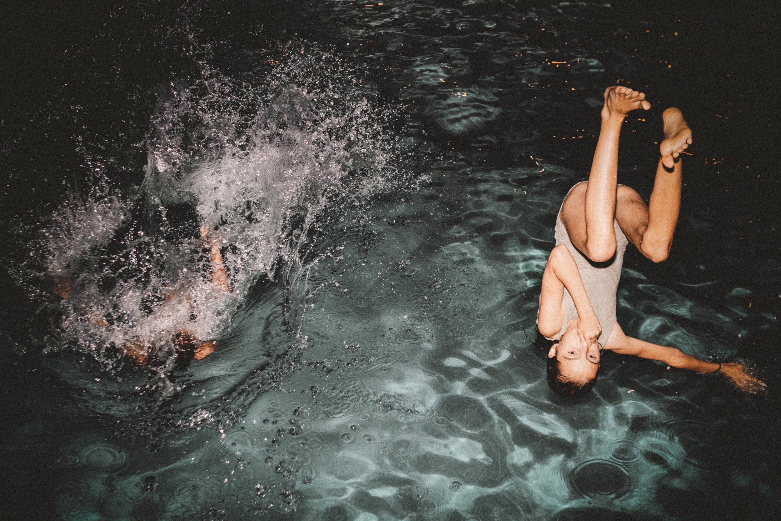 Two children playing in a dark body of water at night, one falling in and splashing water, the other lying on their back with legs in the air.