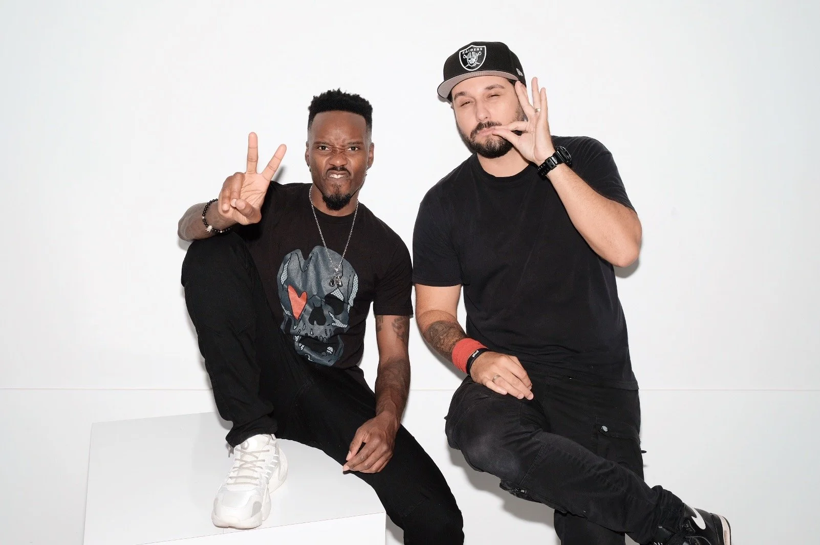 Two men sitting on a white bench against a white background, making peace and hand signs, one wearing a black t-shirt with a skull graphic, and the other wearing a black t-shirt and a black cap with a sports team logo.