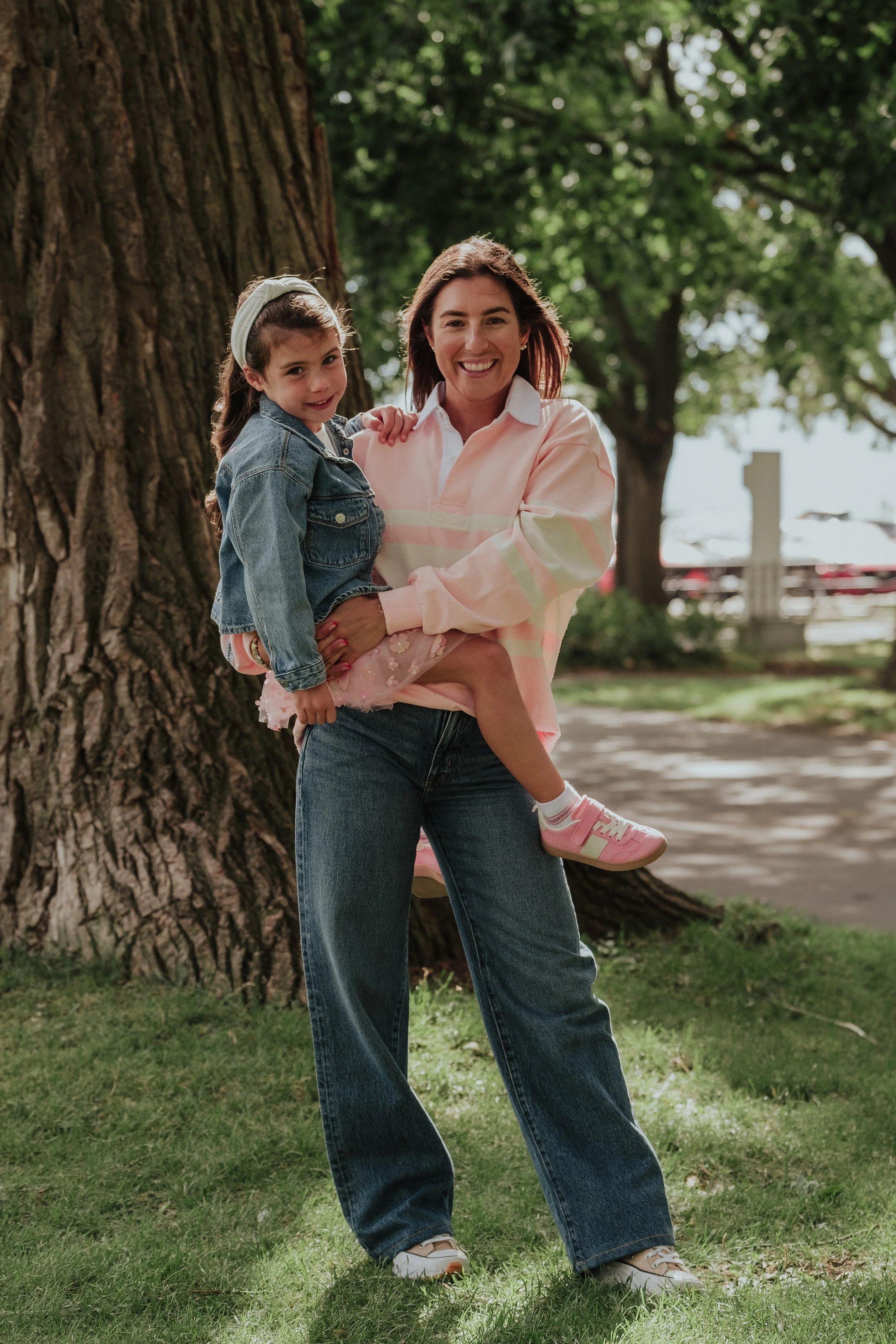A woman holding a young girl in front of a large tree in a park, both smiling at the camera. The woman is wearing a pink and white hoodie, jeans, and sneakers; the girl is wearing a denim jacket, a pink dress, and sneakers.