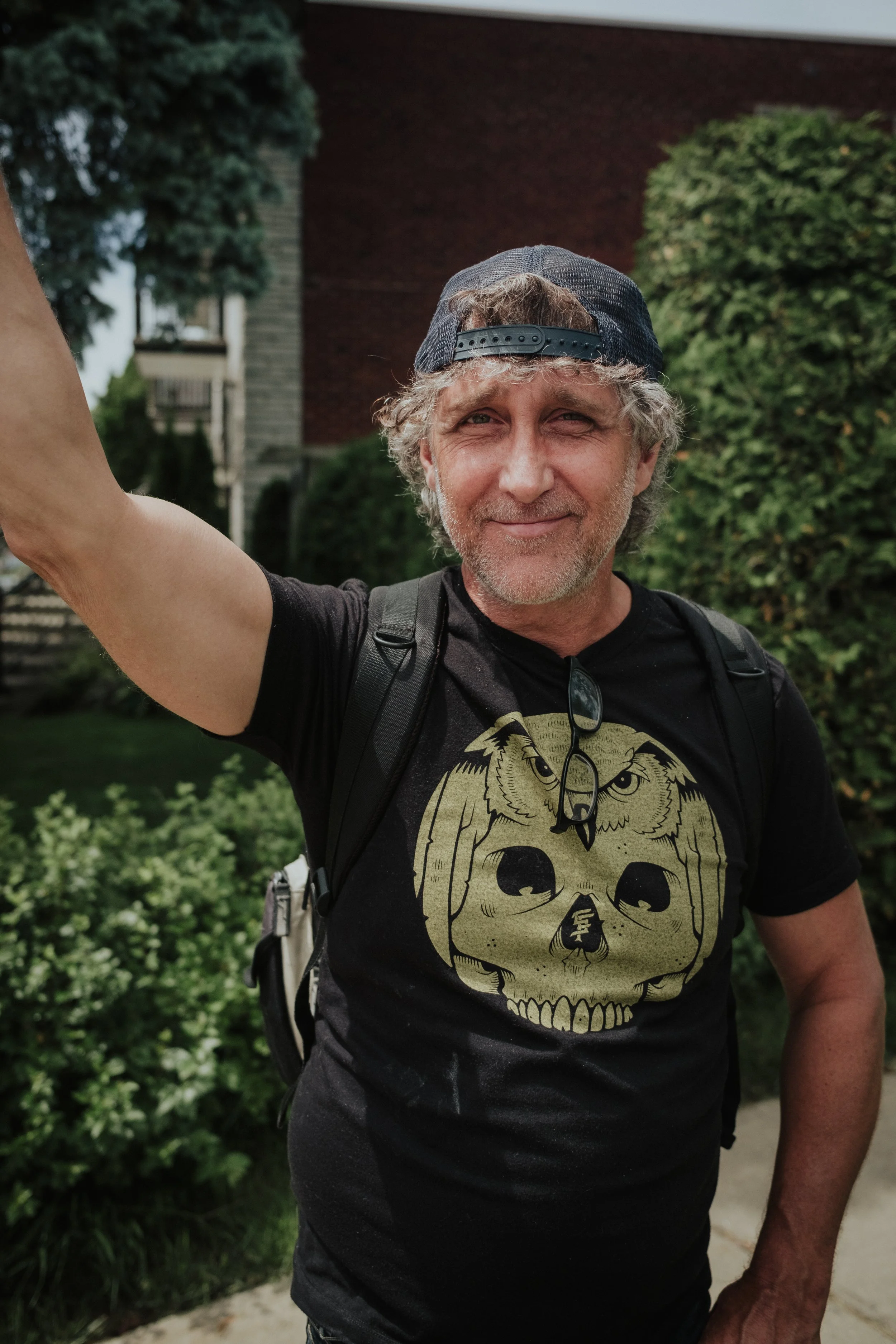A middle-aged man taking a selfie outdoors, wearing a black T-shirt with a skull and owl graphic, a backward baseball cap, glasses hanging on his shirt, and a backpack, standing in front of green bushes and trees.