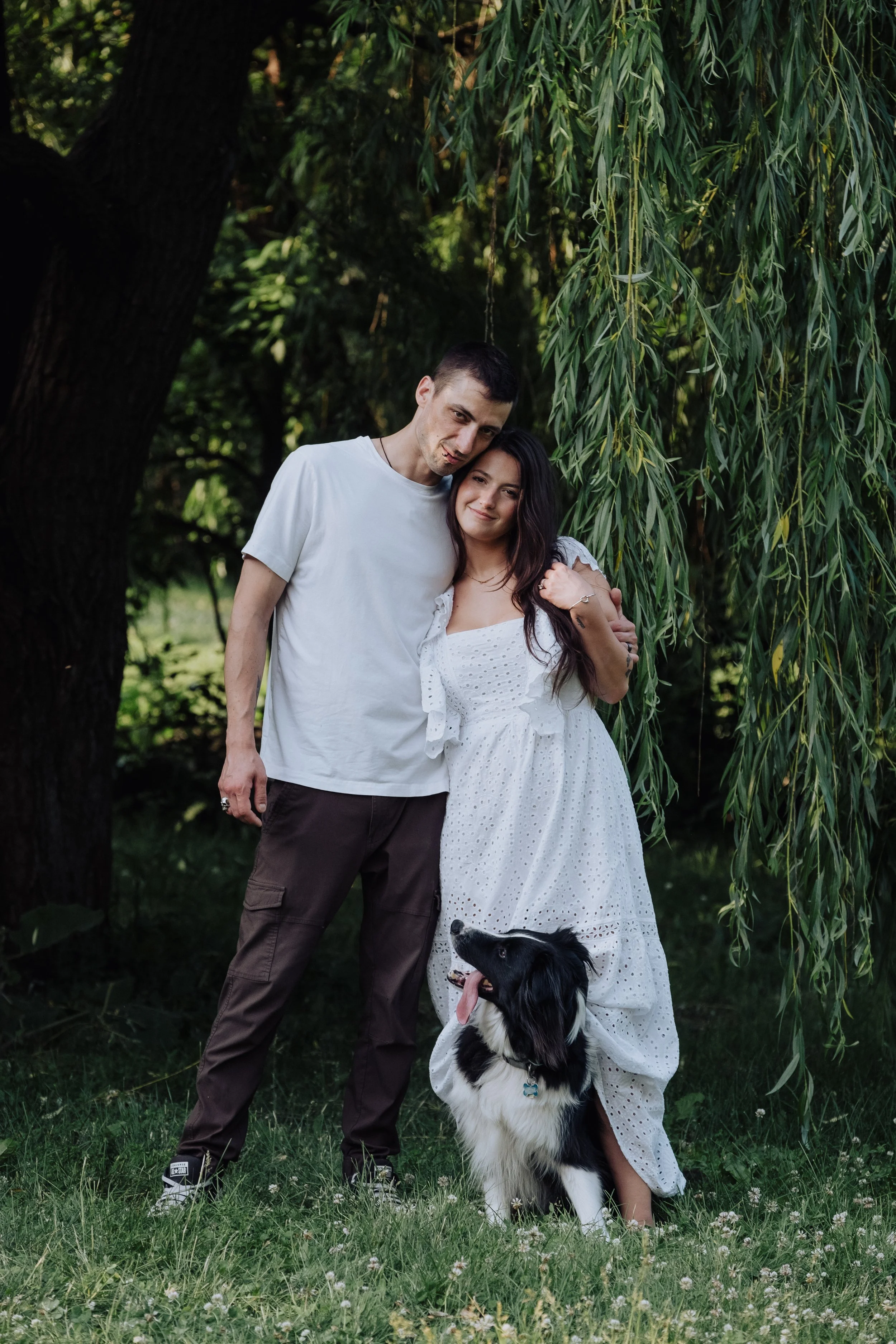 A young couple embracing outdoors with their black and white dog, under a hanging tree with green foliage.