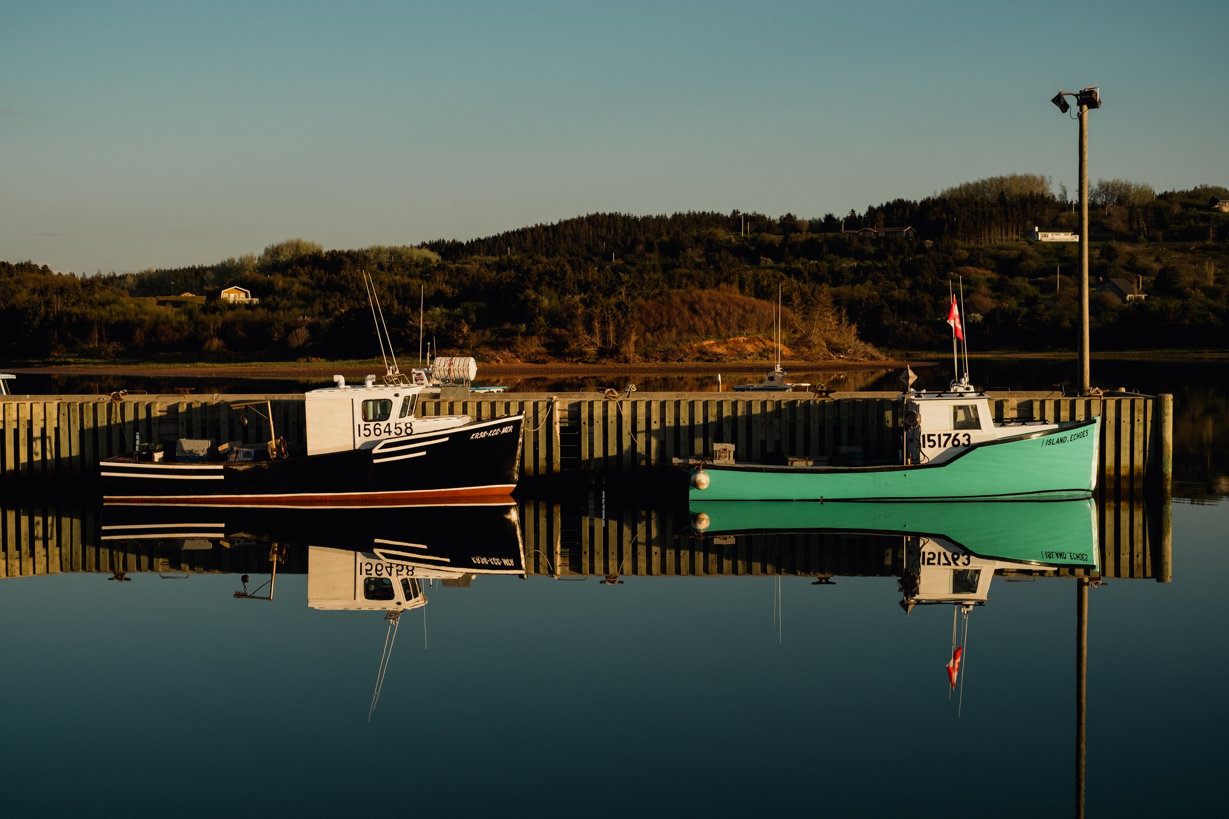 Two boats docked at a wooden pier on a calm body of water, reflecting their images, with a hilly landscape and houses in the background during golden hour.