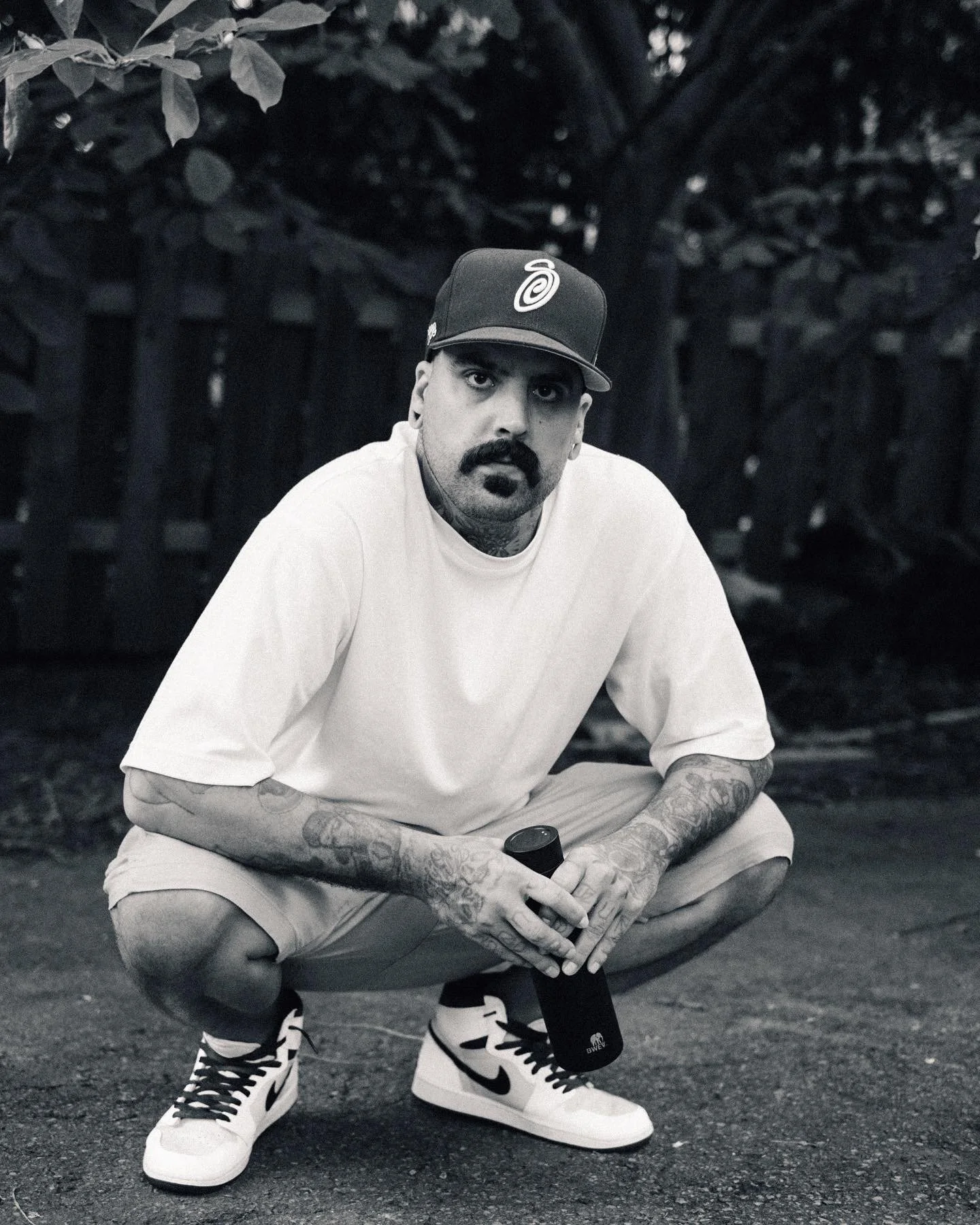 A man with tattoos, a mustache, and a beard, crouching outdoors at night, holding a water bottle, wearing a white t-shirt, grey shorts, white and black Nike sneakers, and a baseball cap with a logo.