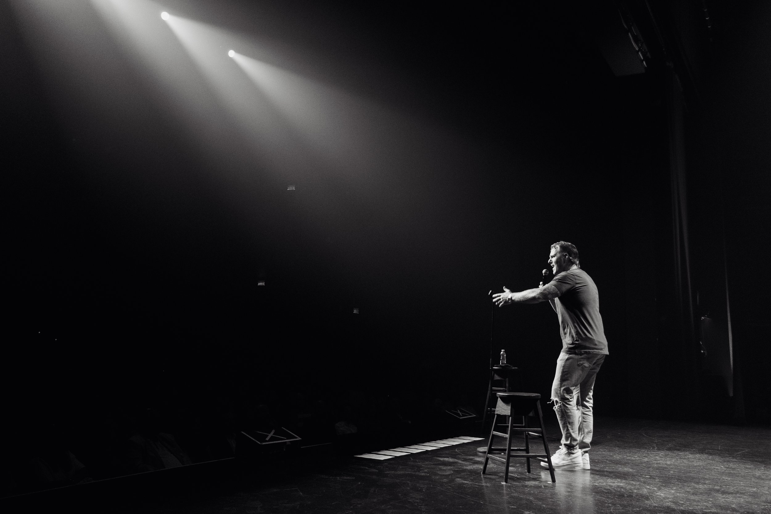 A comedian stands on a stage with a microphone, gesturing with his right hand, under bright spotlights, in a darkened auditorium.