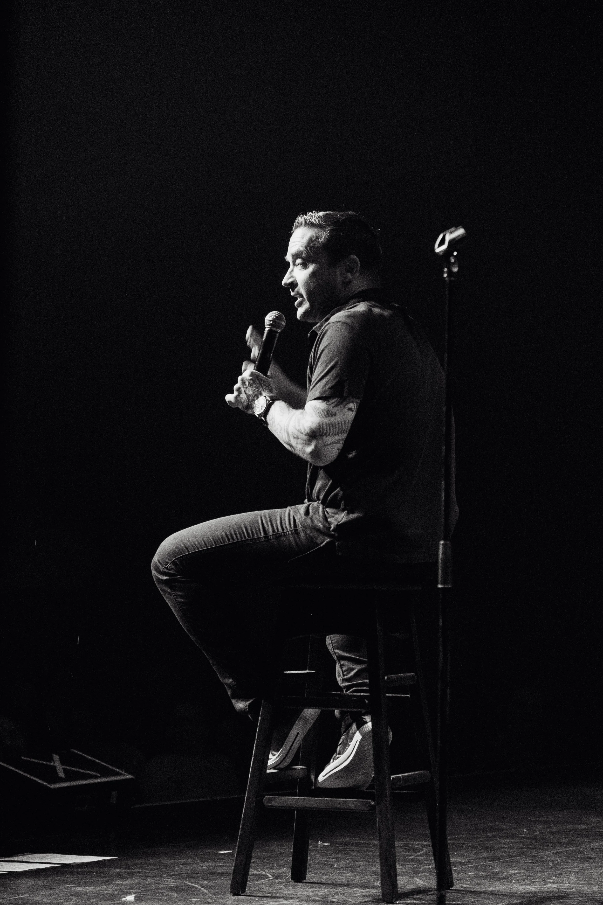 A man sitting on a high stool holds a microphone, engaged in speaking or performing on stage, with a dark background.