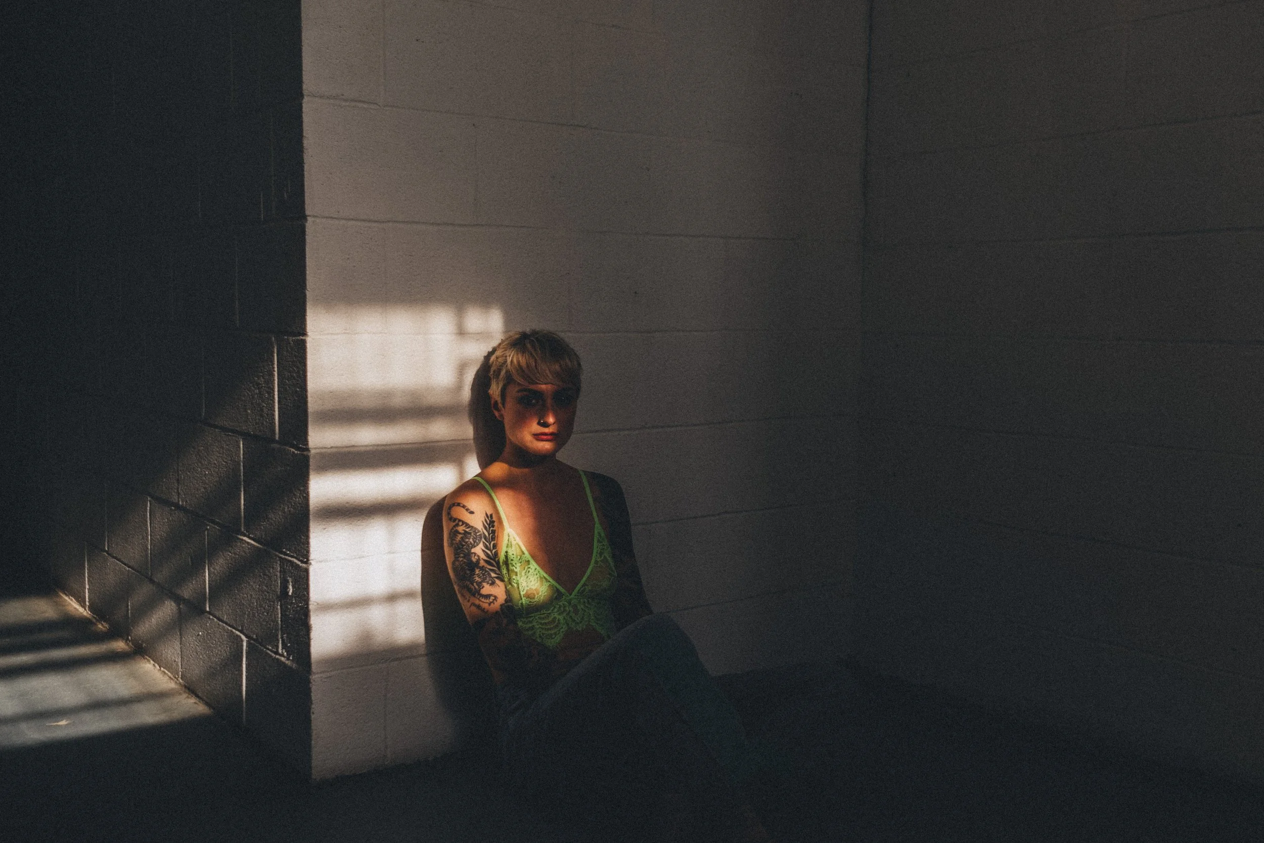 A woman with short hair and tattoos on her arm sits on the floor in a dimly lit room with concrete walls, illuminated by sunlight through blinds casting shadows.