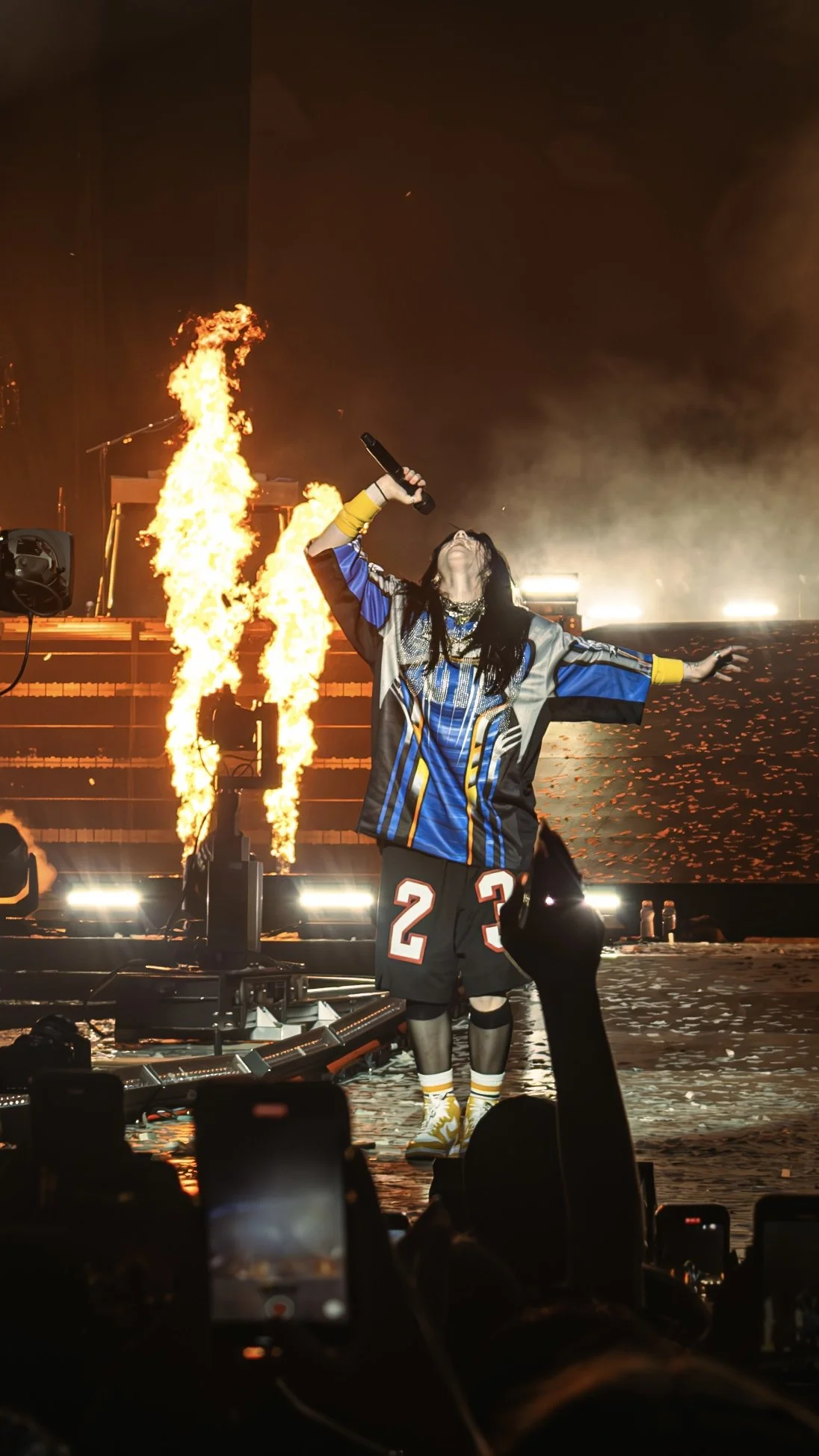 A performer on stage wearing a colorful jersey, black shorts with the number 23, knee-high socks, and athletic shoes, singing into a microphone with flames and bright lights in the background, and audience members capturing the moment.