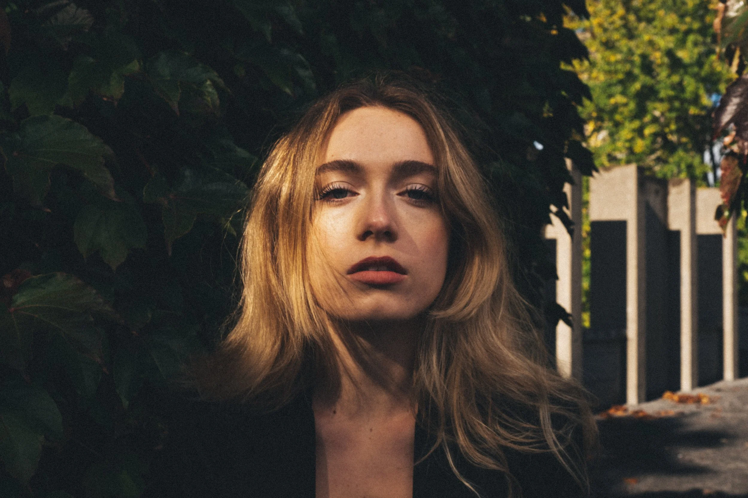 A young woman with long, wavy blonde hair looking at the camera with a serious expression, standing near dark green leafy bushes on a sidewalk with trees and a fence in the background during daytime.
