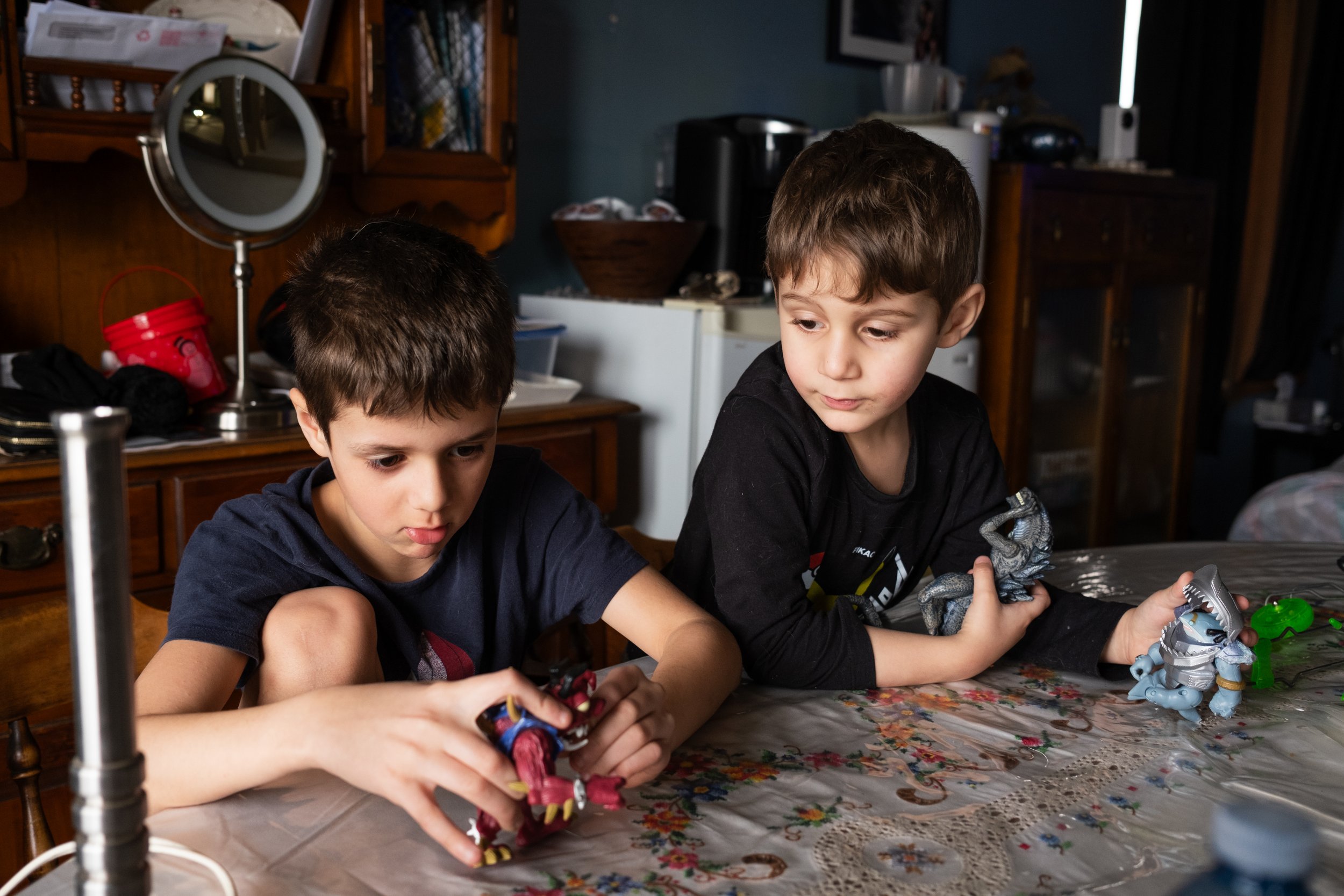 Two boys sitting at a dining table playing with toy dinosaur figures in a room with wooden furniture and household items in the background.