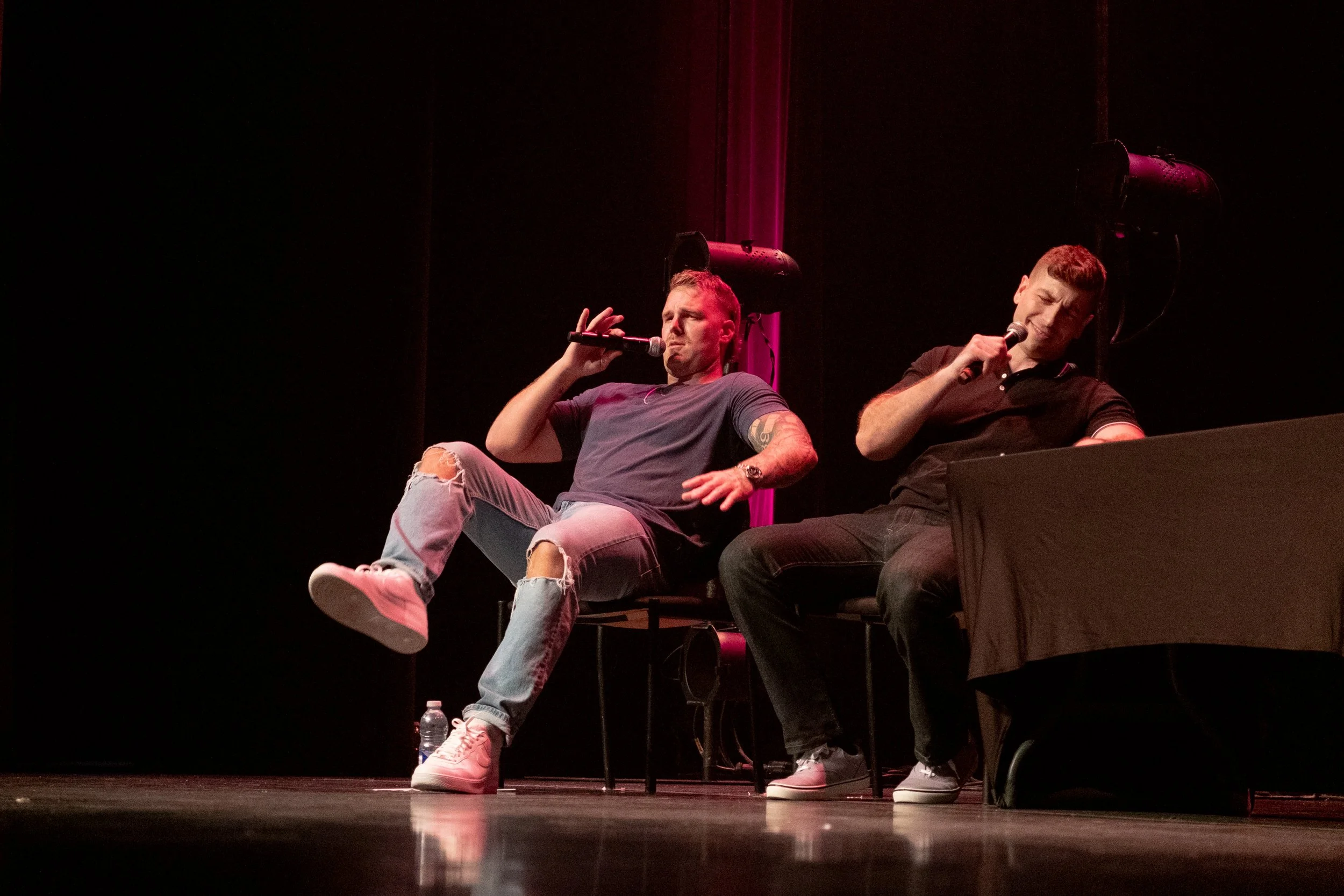 Two men sitting on chairs on a stage, each holding a microphone. The man on the left is wearing a gray T-shirt, ripped jeans, and white sneakers, with one leg crossed over the other. The man on the right is wearing a black polo shirt and jeans, sitti