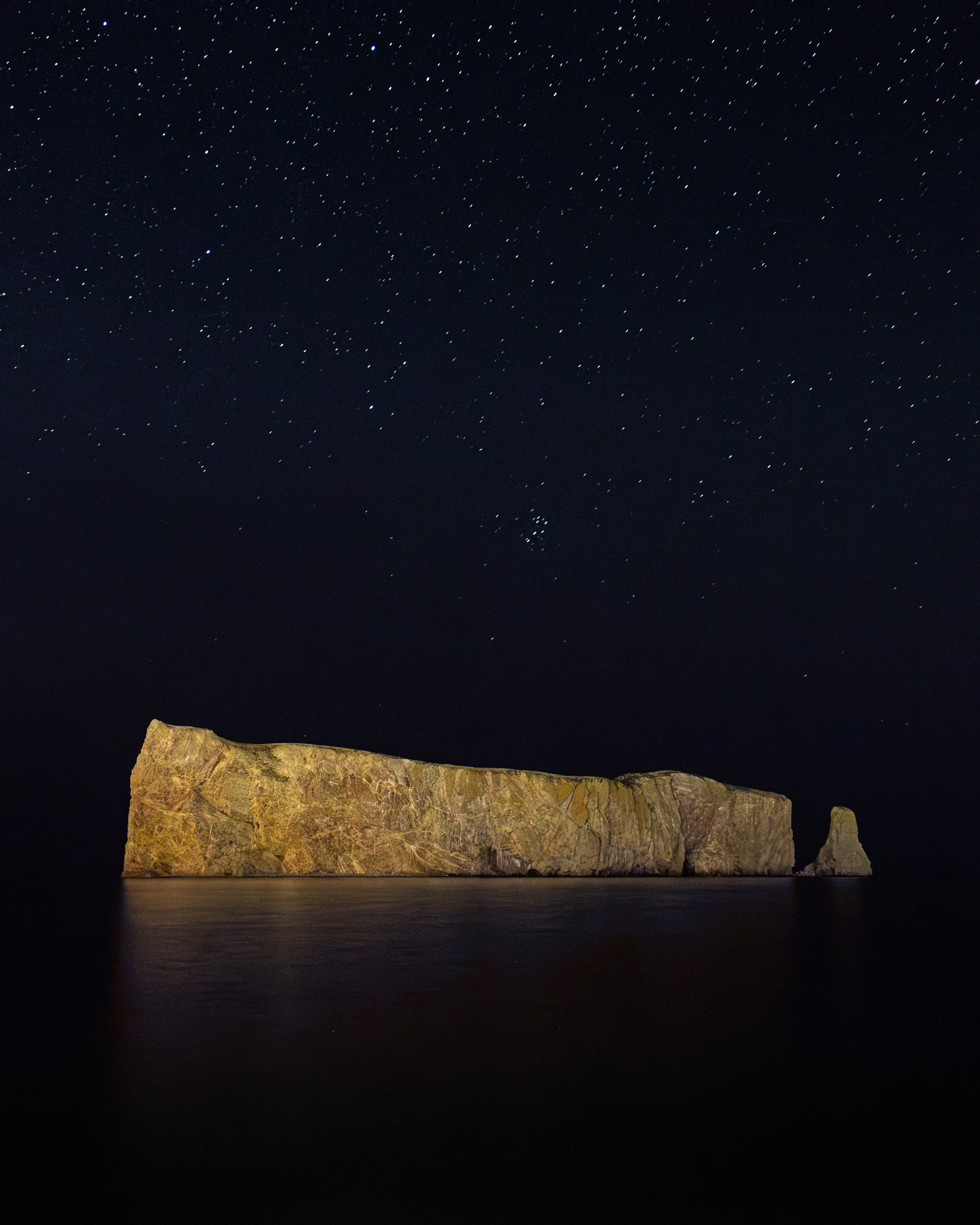 Nighttime image of a large rocky island or formation surrounded by water, with a starry sky overhead.