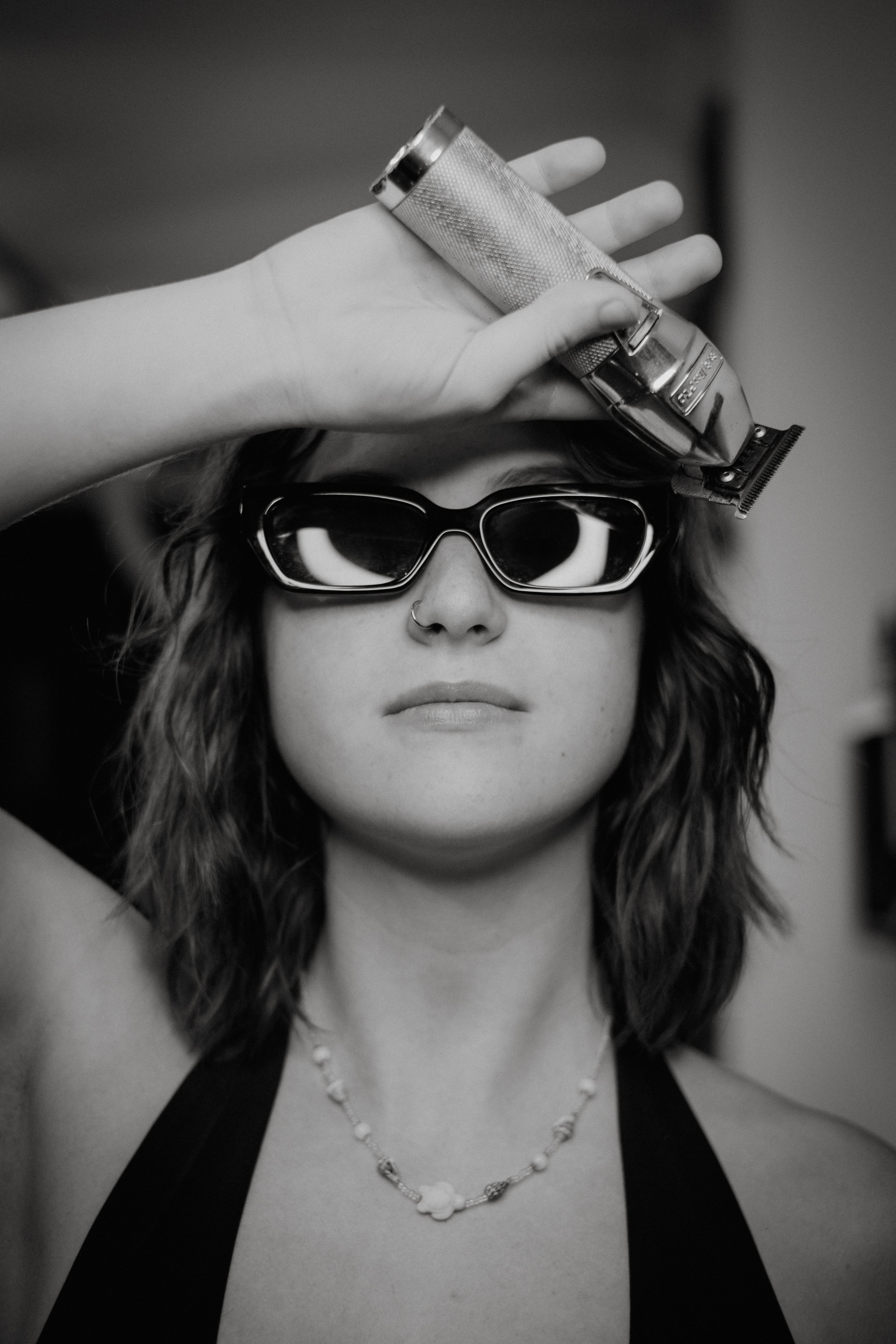 A woman with wavy hair wearing sunglasses and a necklace, holding a hair clipper above her head as if preparing to cut her hair.