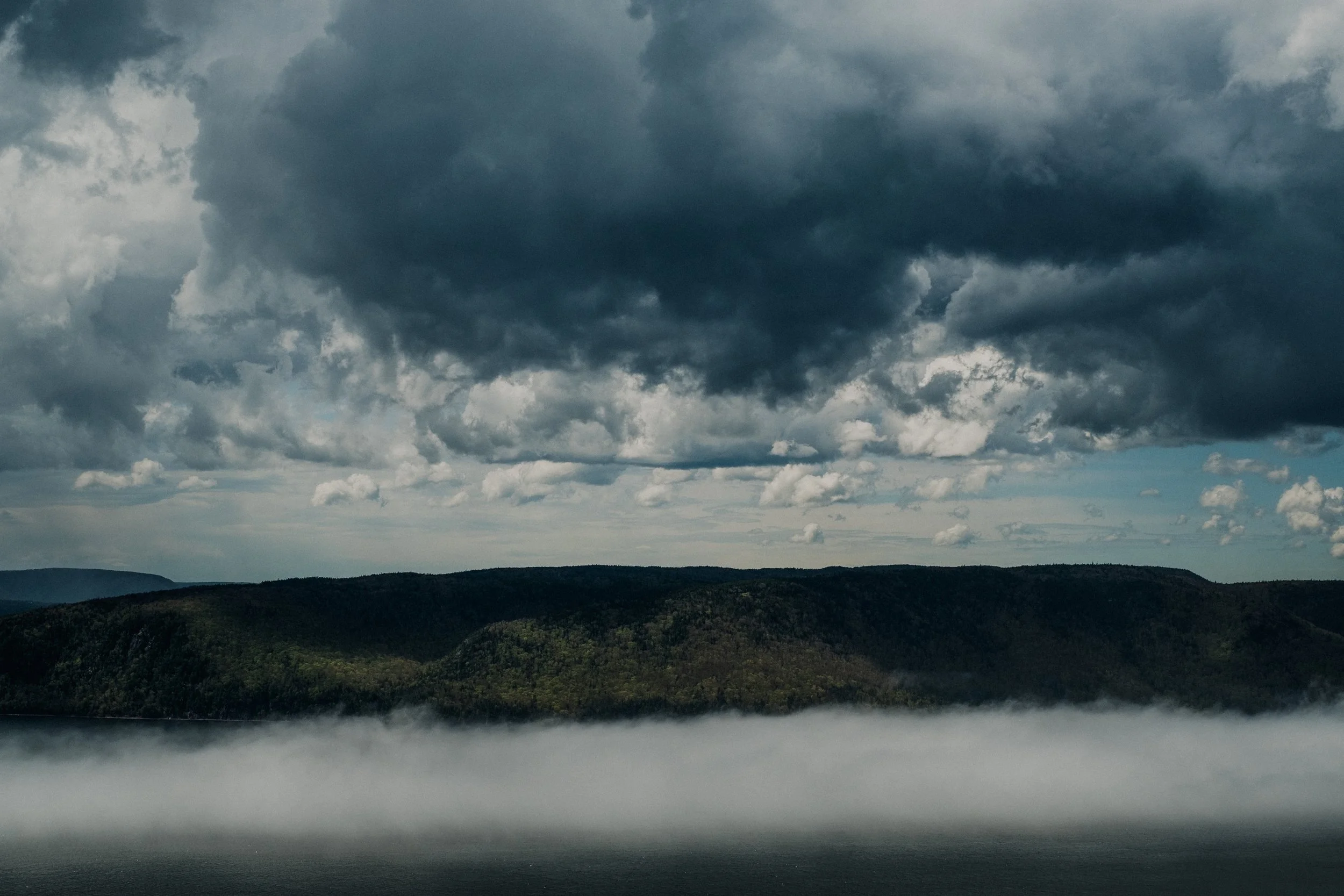 Dark storm clouds over a mountain range with fog near a body of water in the foreground.