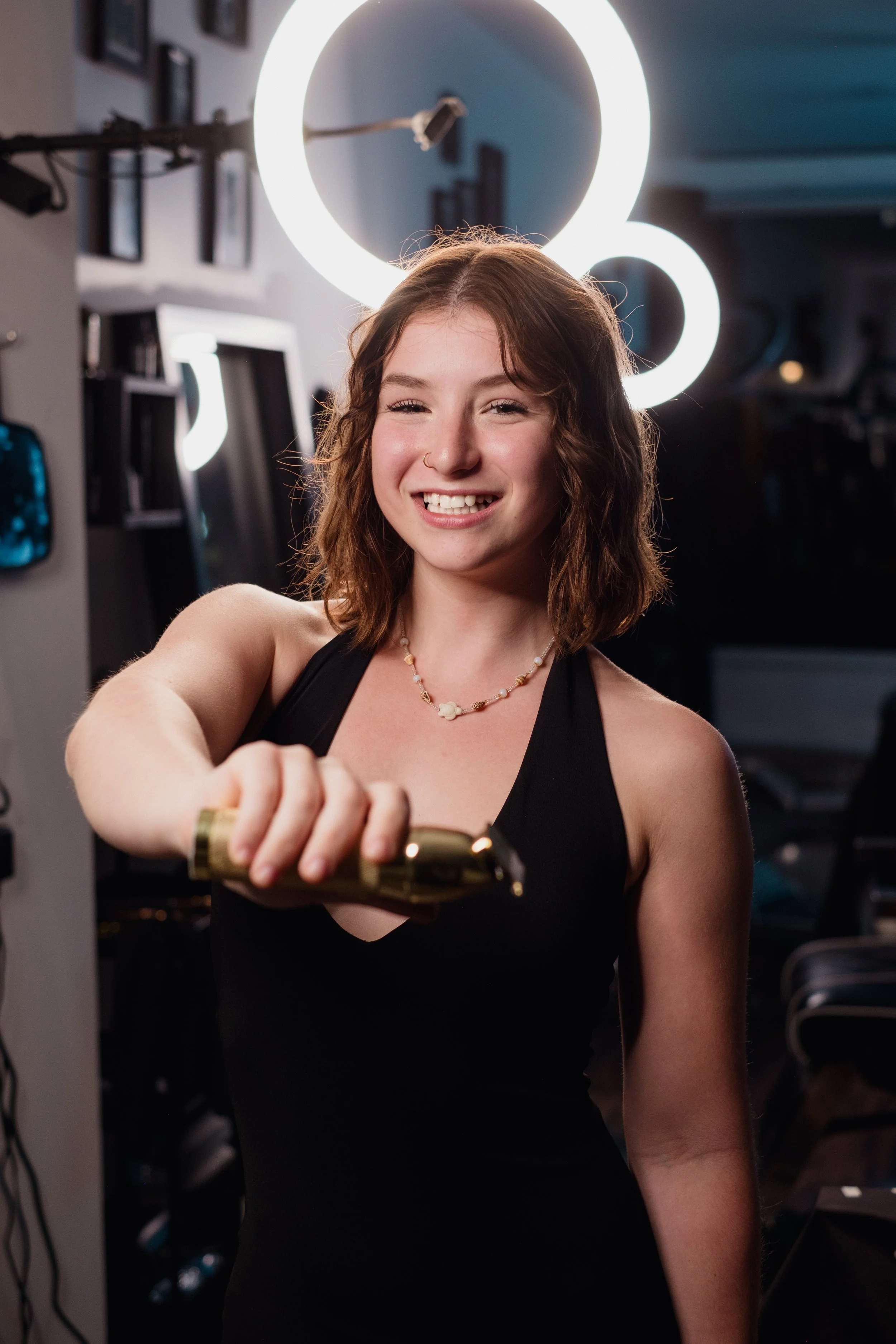 A woman with shoulder-length wavy hair is smiling and holding a hair clipper towards the camera.
