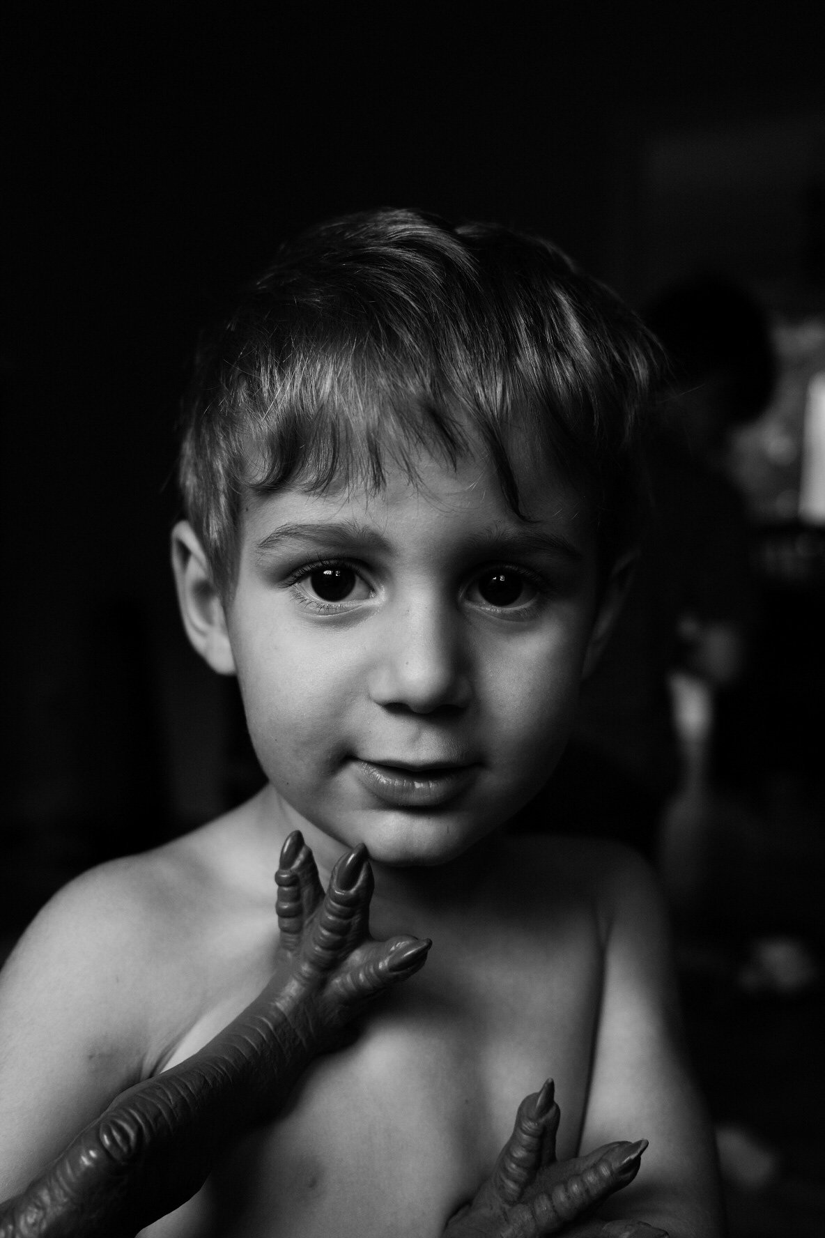 A young boy with short hair looks directly at the camera, with wide eyes and a slight smile, as a person with twisted fingers touches his shoulder.