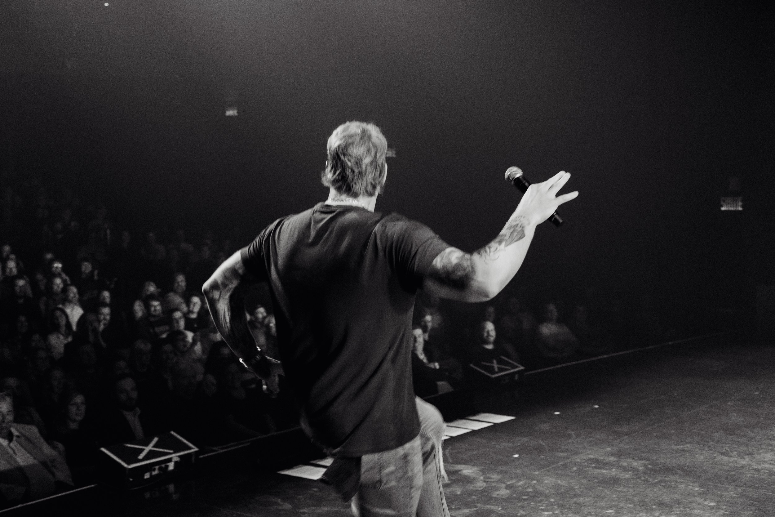 A male performer with tattoos on his right arm, holding a microphone in his right hand, is on stage facing an audience, gesturing with his left hand during a live performance in a dark theater.