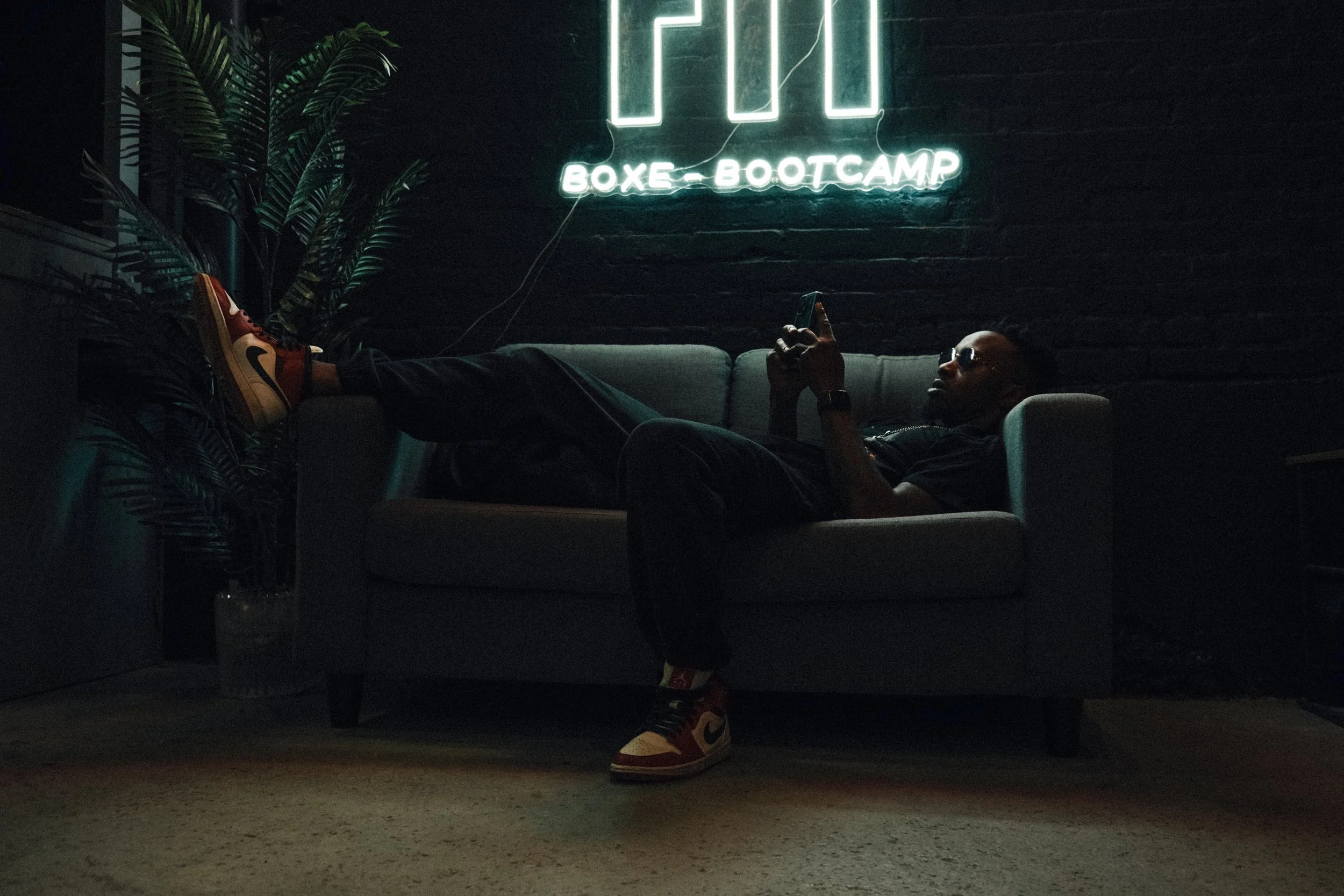 A man relaxing on a gray sofa in a dark room with a neon sign that reads 'BOXE BOOTCAMP' on a black brick wall behind him. He is lying back with his feet up, wearing sunglasses, black clothing, and sneakers, and is looking at his phone. There is a la