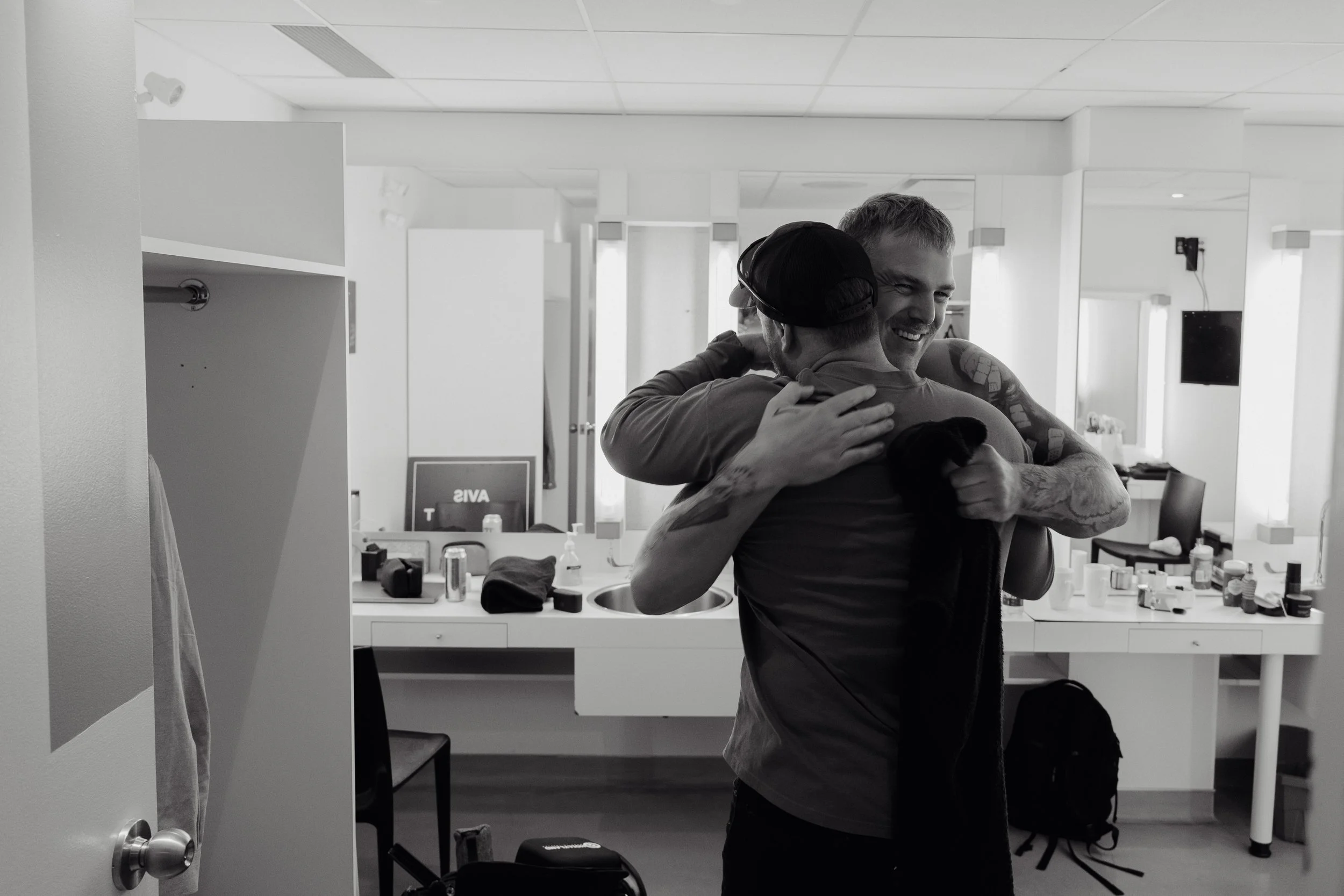 Two men hugging in a bathroom mirror, smiling and embracing each other.