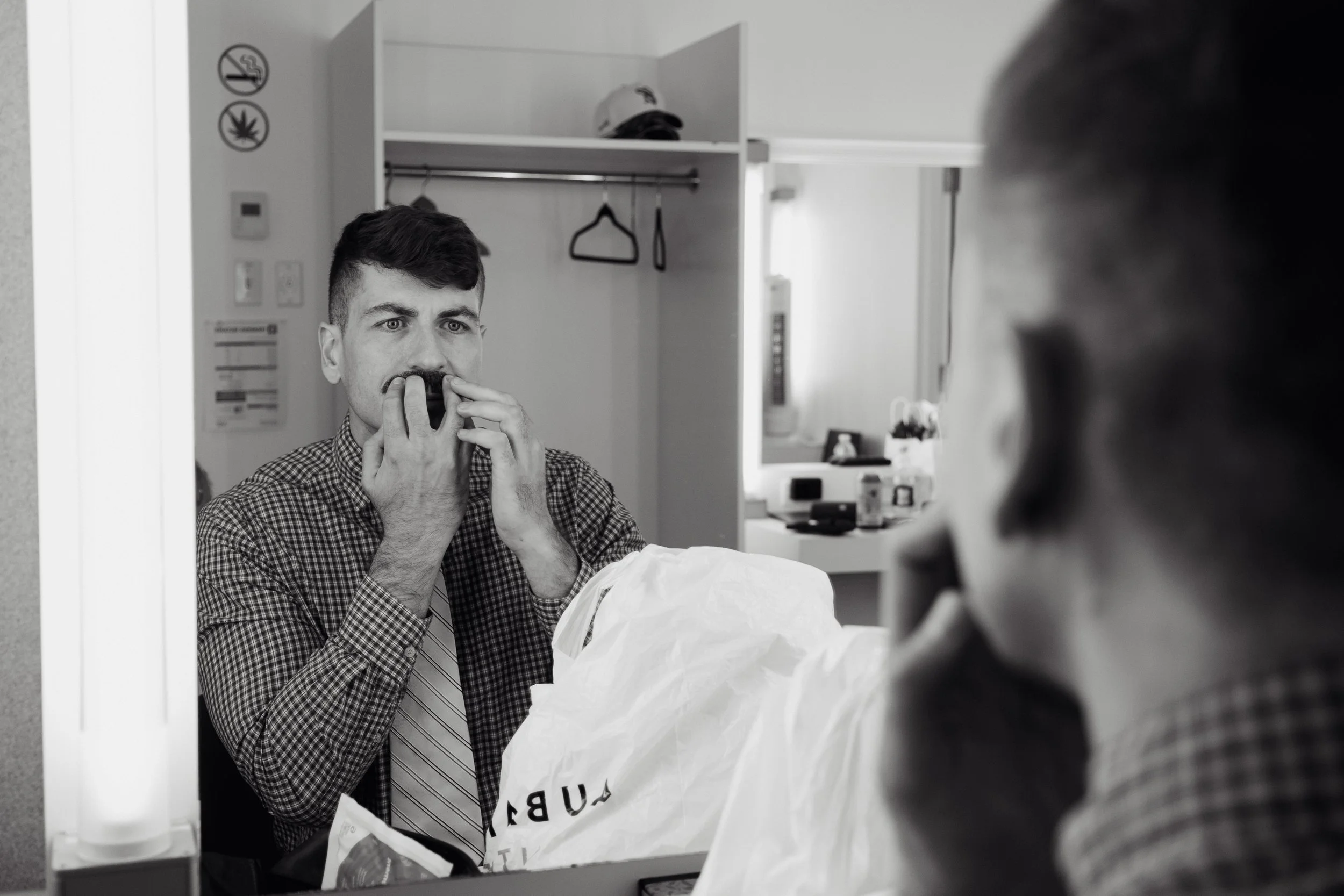 A man looking at himself in a mirror while holding a mustache prop to his face, with a hospital room in the background.