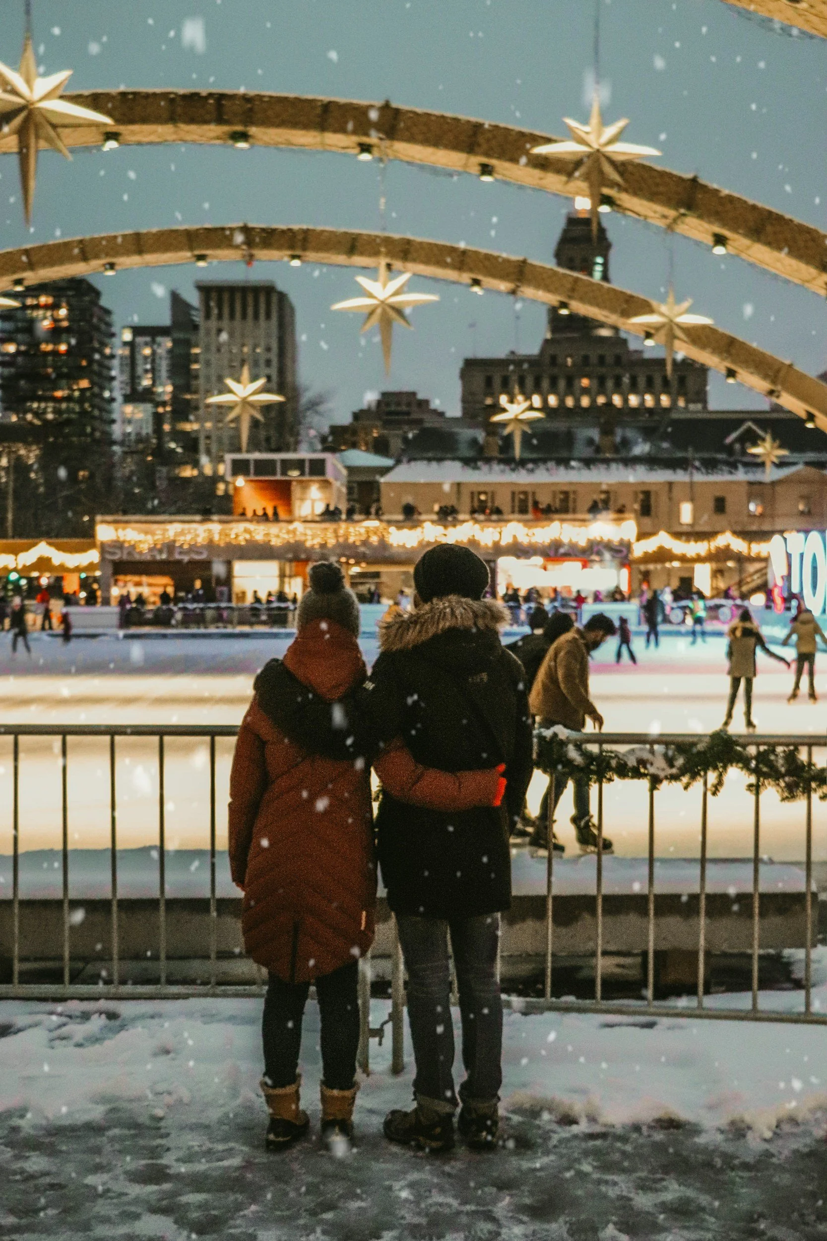 Couple standing together at a winter ice rink beneath festive Christmas lights and decorations