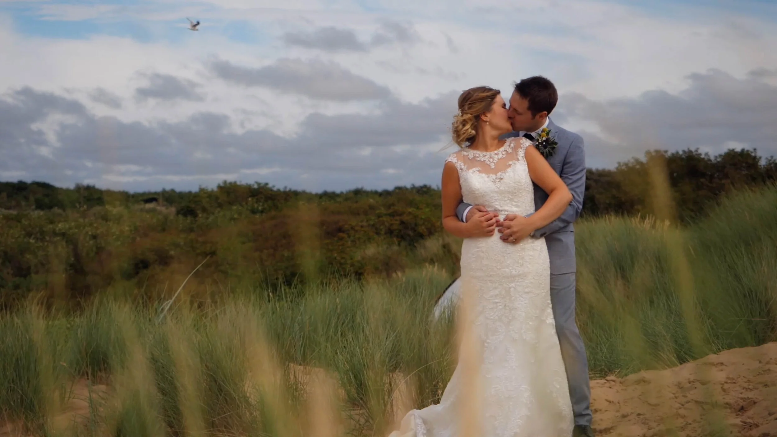 Finn + Emma share a kiss on Formby Beach, Liverpool. The two are kissing amongst the sand dunes, as a bird flies above in the cloudy sky.