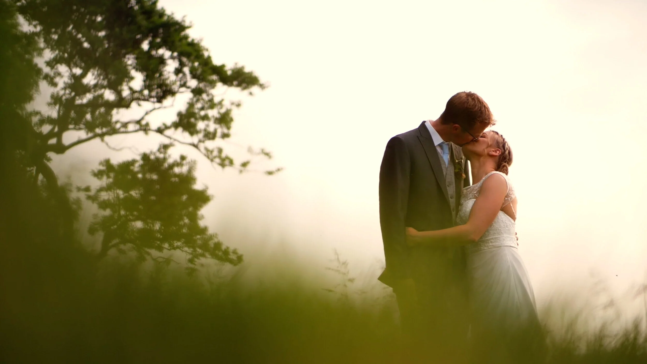 David and Lisa share a kiss amidst an overgrown field. The sun setting behind them, and the trees waving in the wind.