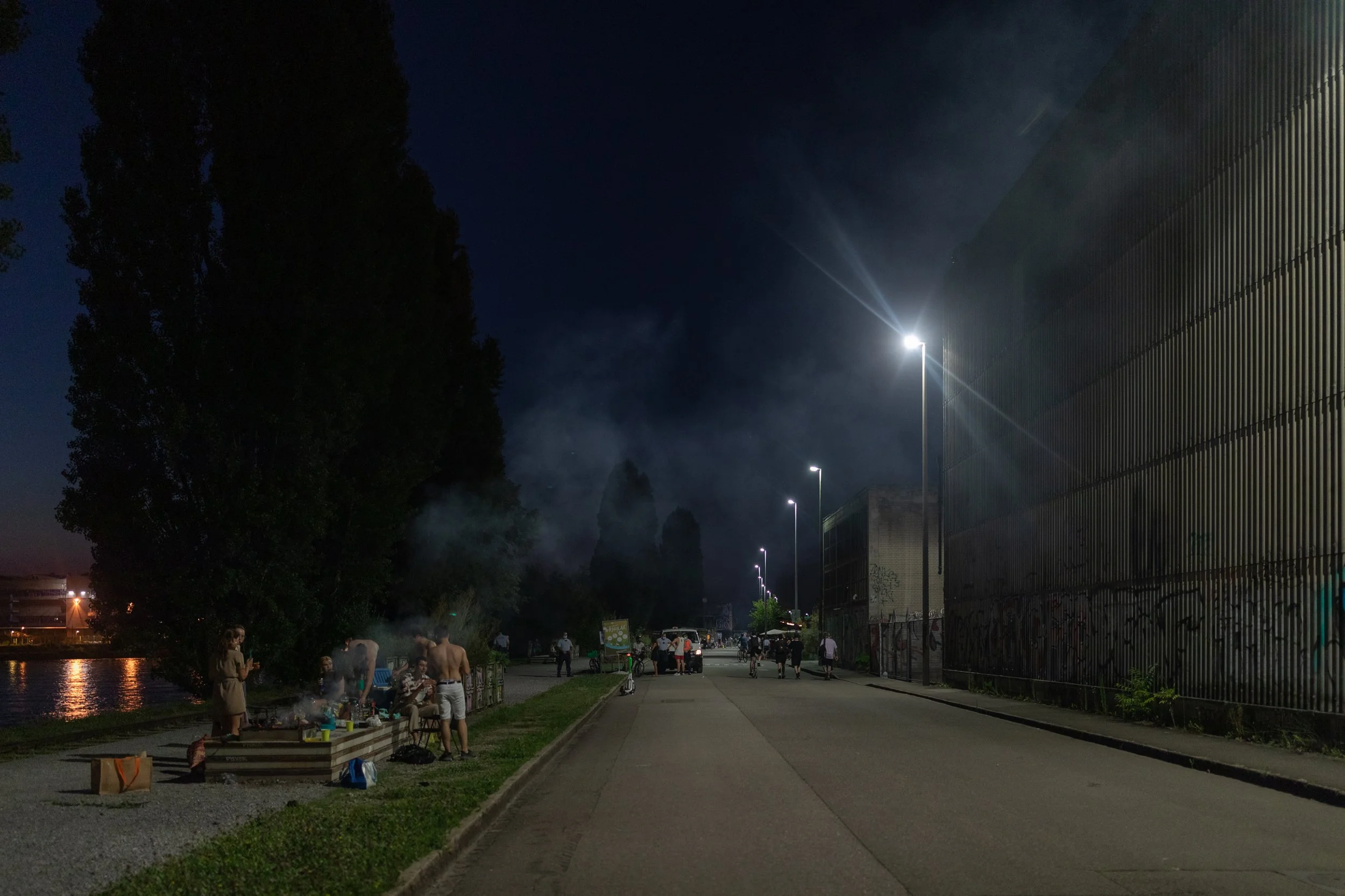 Menschen sitzen und stehen am Flussufer bei Nacht, während im Hintergrund eine große moderne Gebäudefront mit vertikalen Linien steht und Straßenlaternen leuchten.