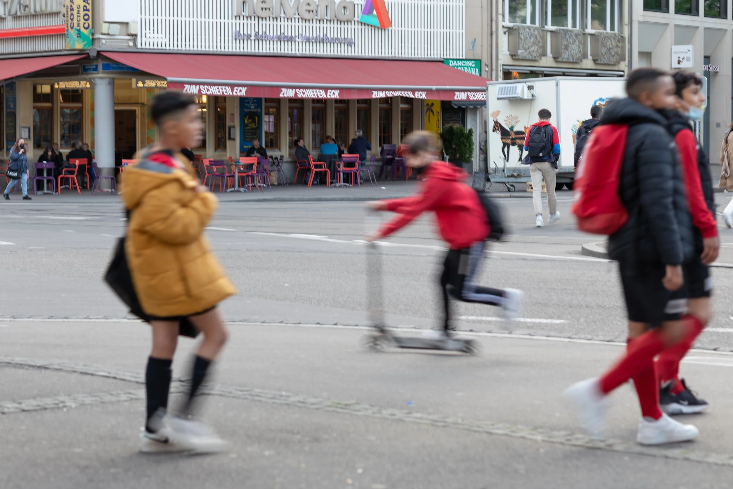 Menschen laufen über eine Straßenkreuzung vor einem Restaurant mit aufgestellter Markise. Einige sitzen draußen, während mehrere Personen vorbeigehen, darunter Kinder in roten und schwarzen Jacken.