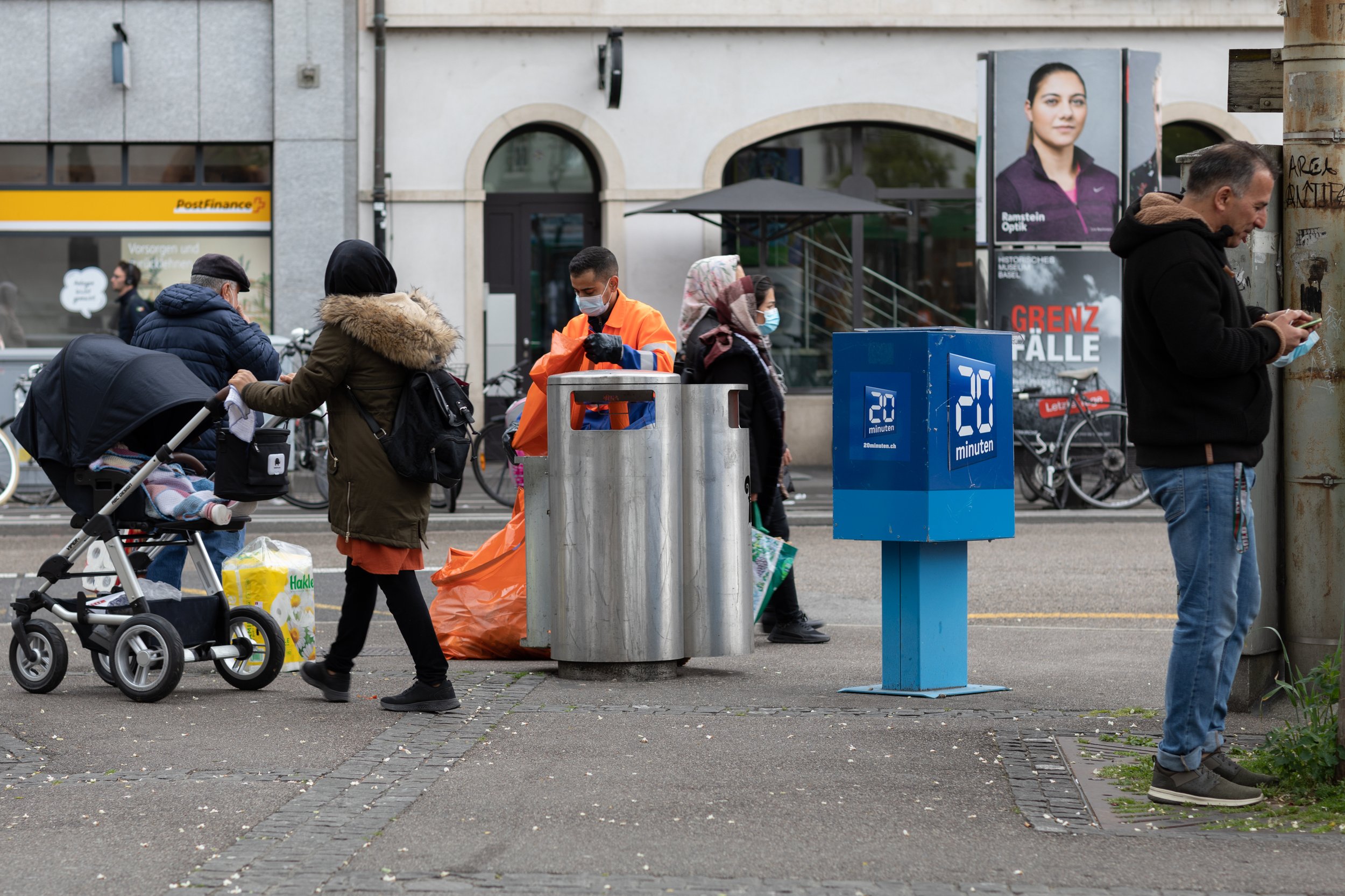 Menschen stehen an einer Straßensperre, einige tragen Masken, eine Person in einer orangefarbenen Jacke arbeitet mit einem Scanner, verschiedene Fahrradständer und ein digitaler Zähler sind sichtbar, im Hintergrund sind Werbetafeln und ein Eingang zu