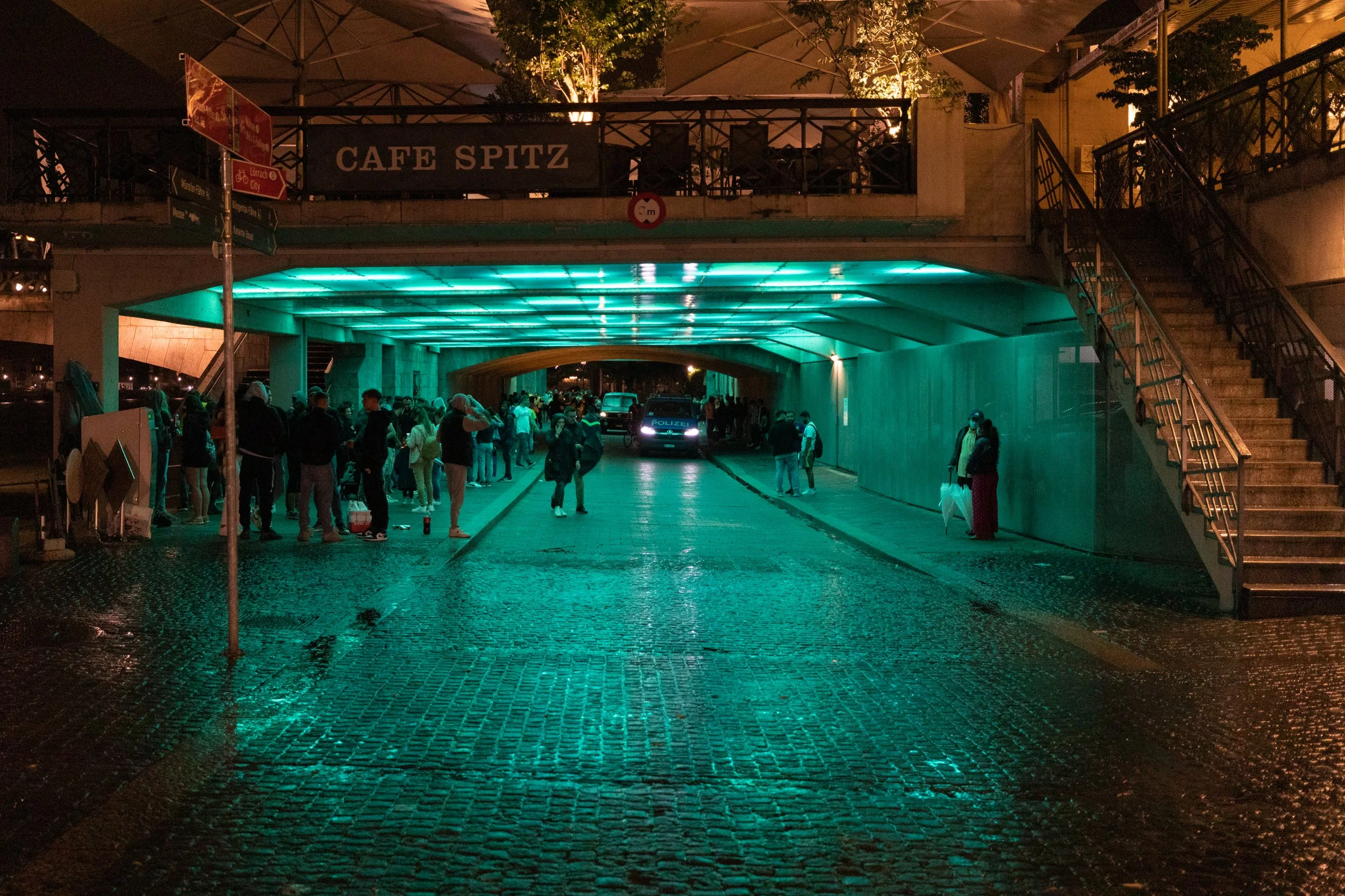 Menschen versammeln sich in einem Tunnel unter einer Brücke, die mit grünen Lichtern beleuchtet ist, Frauen und Männer stehen in Gruppen, einige unterhalten sich, Polizei im Hintergrund, im oberen Bereich ein Café mit Tischen und Stühlen auf einer Te