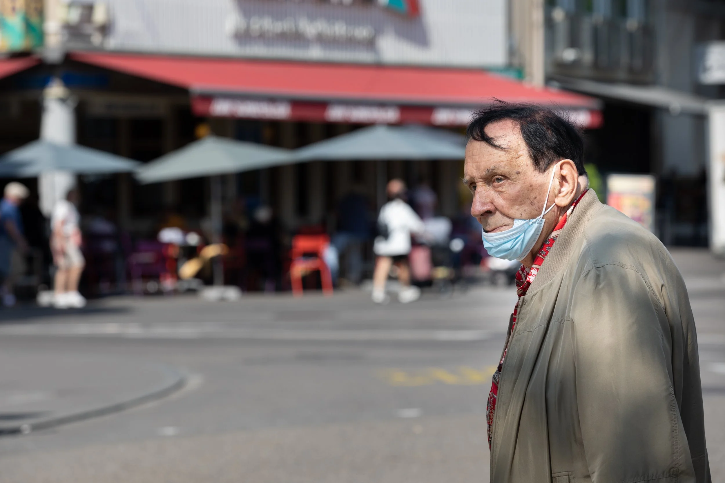 Ein älterer Mann mit dunklem Haar trägt eine blaue Schutzmaske, während er die Straße überquert. Im Hintergrund sind Straßen Cafés mit Tischen und Stühlen, einige Menschen laufen vorbei, und es gibt eine Geschäftsfront an einer belebten Stadtstraße.