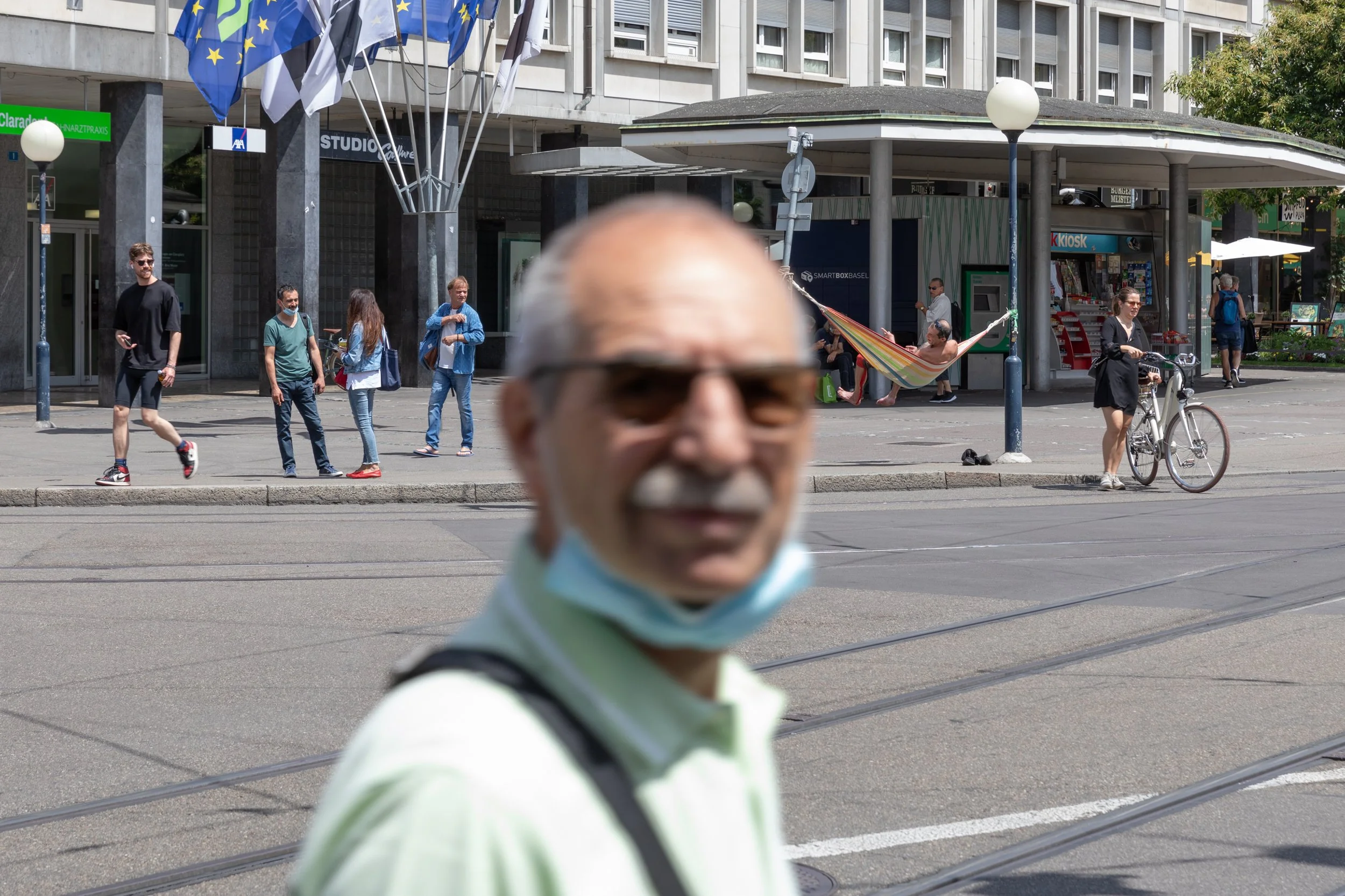 Ein älterer Mann mit Sonnenbrille und Maske, der im Vordergrund unscharf ist. Im Hintergrund sind mehrere Menschen auf einer Stadtstraße, einige laufen, andere sitzen in einem Hängematte, eine Person fährt Fahrrad. Es gibt Flaggen, eine Kiosk-Station