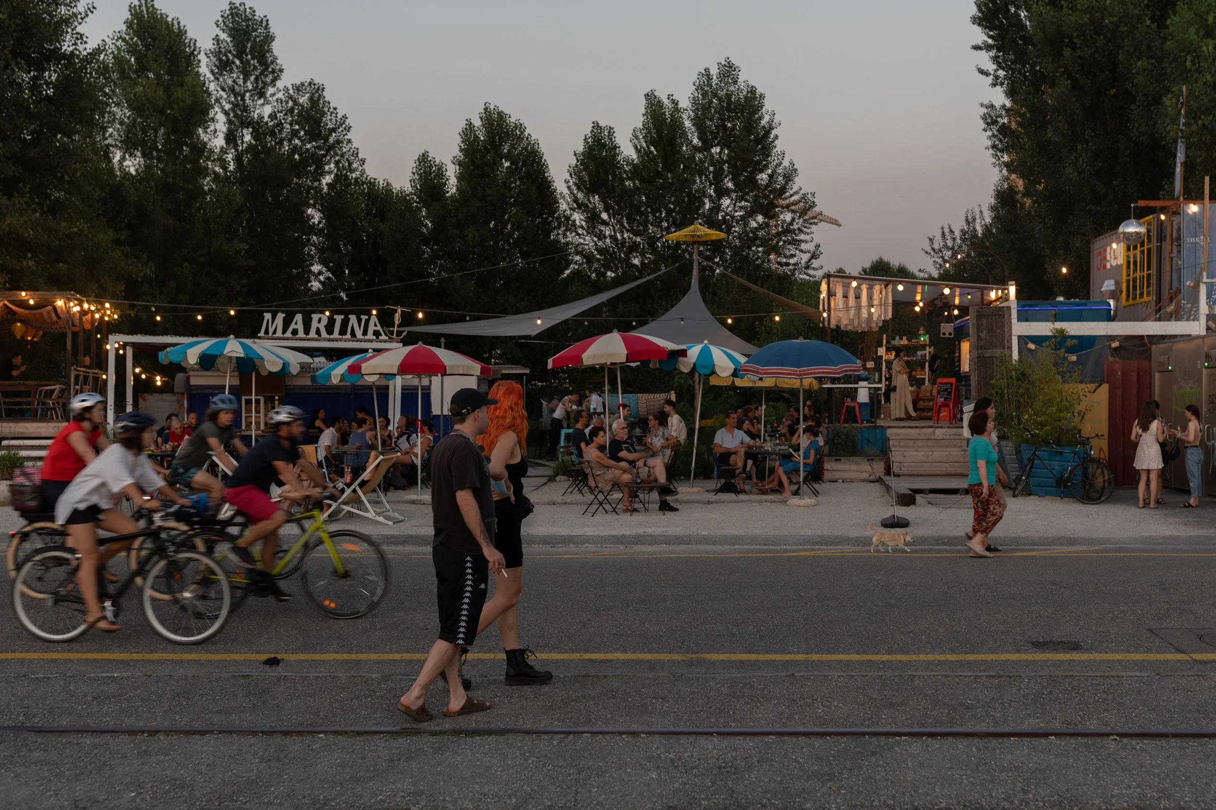 Menschen radeln, stehen und spazieren an einer belebten Strandpromenade mit Tisches, Sonnenschirmen und Lichterketten bei Sonnenuntergang. Im Hintergrund sind Bäume, ein Kran und eine gastronomische Anlage sichtbar.
