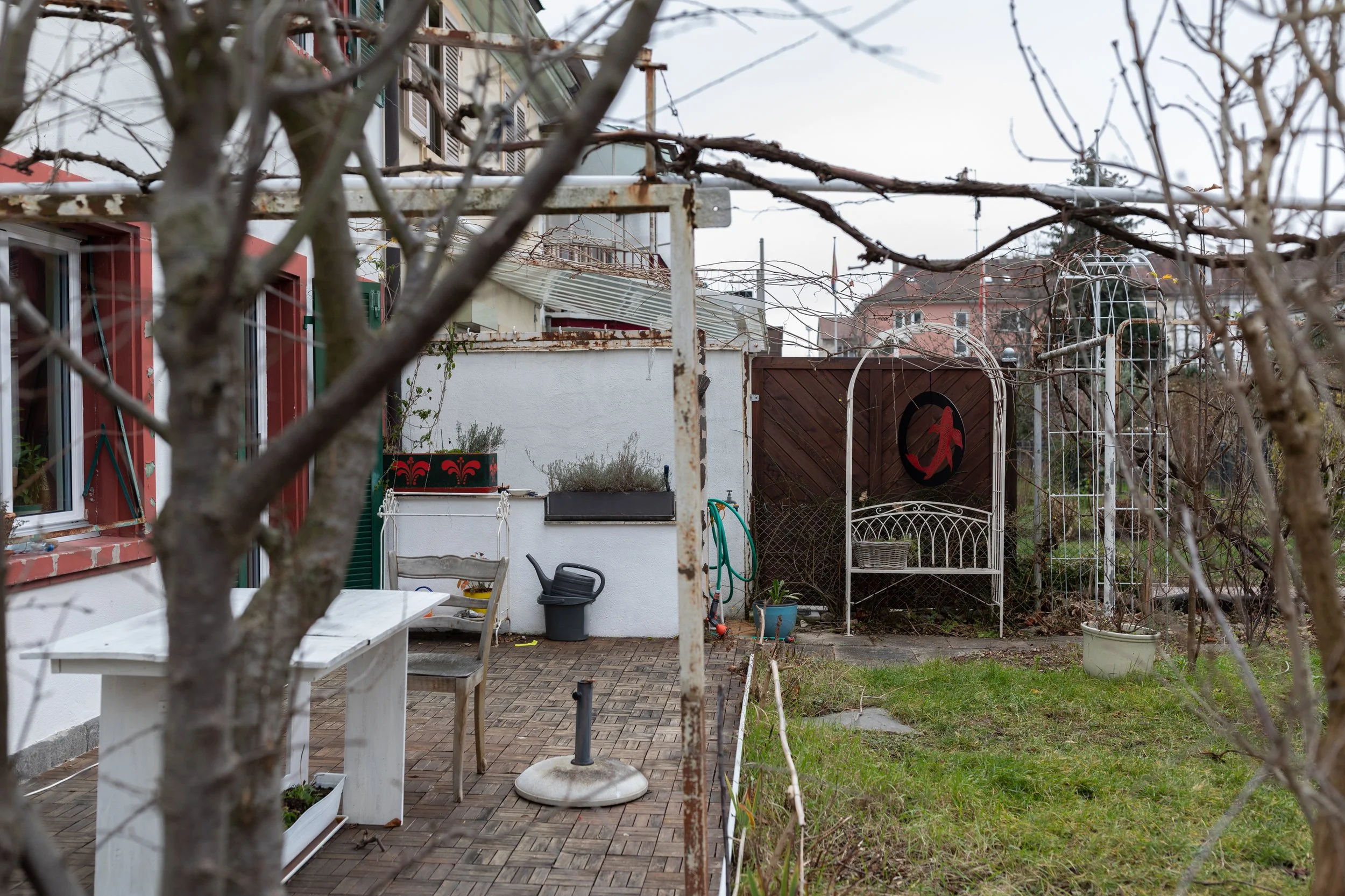 Ein Garten mit Baum, Gartenmöbeln, Pflanzen, Gartenzaun und einem Gebäude im Hintergrund an einem bewölkten Tag.