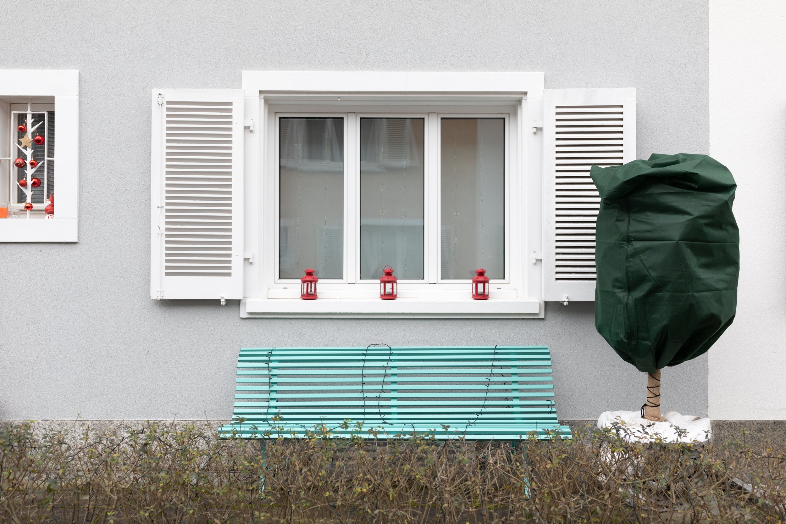 Ein Fenster mit weißen Jalousien und Dekorationen, darunter vier rote Laternen, auf einem weißen Fensterbrett. Im Vordergrund ein Baum mit grünem Blätterdach, der mit einer Laserlichtdekoration versehen ist, und im unteren Bereich ein blauer Gartenst