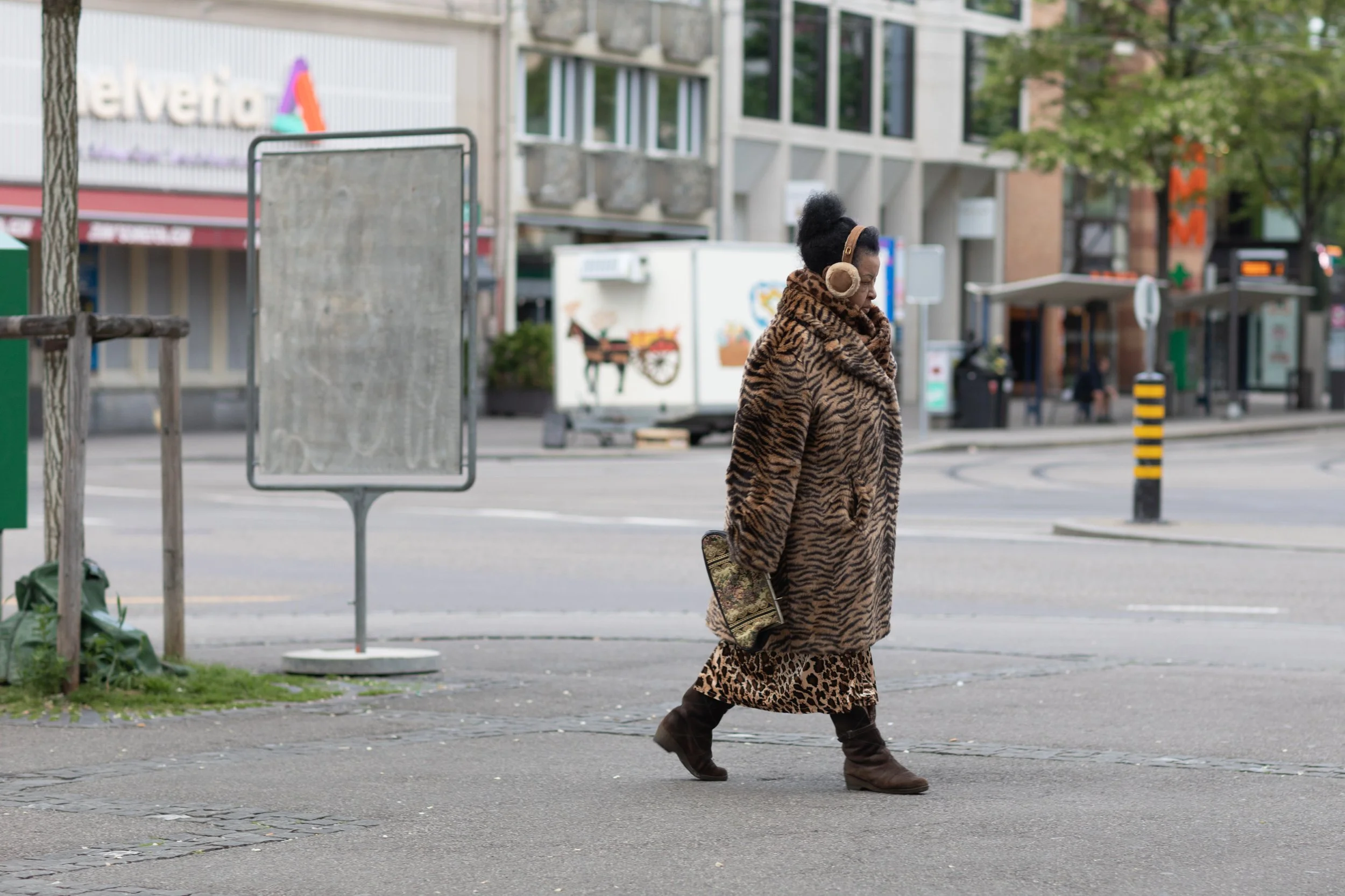 Frau in einem Leopardenmuster Mantel und Boots, die die Straße entlanggeht, trägt Kopfhörer, während sie die Straße überquert, mit städtischer Umgebung im Hintergrund