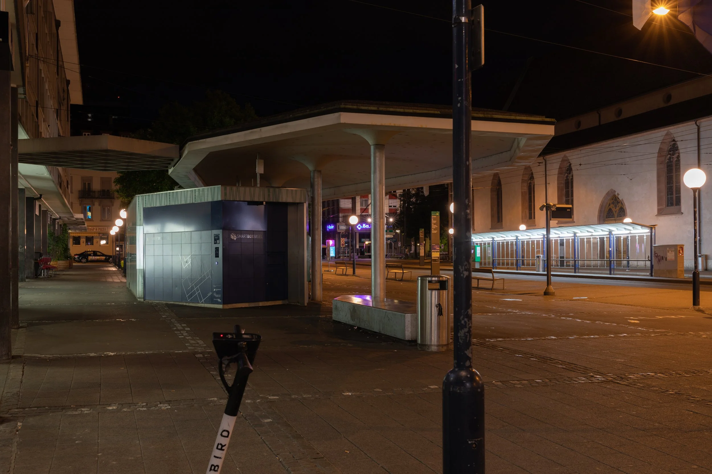 Leerer Stadtplatz bei Nacht mit U-Bahn-Station, Stadtbänken, einer Litfaßsäule, Straßenlaternen und einem modernen Wartehäuschen.