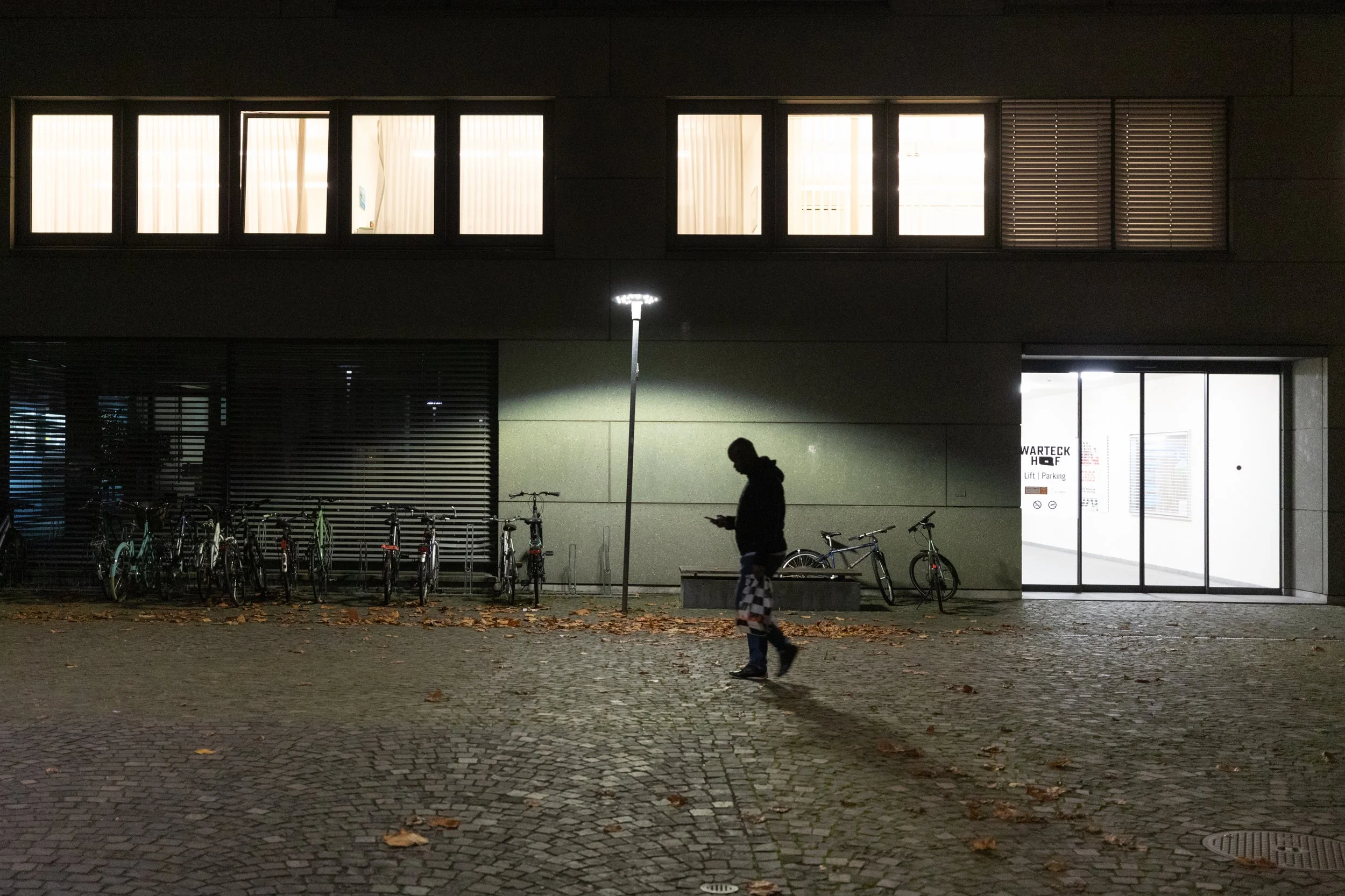 Ein Mann geht nachts an einem beleuchteten Fahrradständer mit mehreren Fahrrädern vorbei, im Hintergrund ein modernes Gebäude mit großen hier beleuchteten Fenstern und verzierten Fensterläden.
