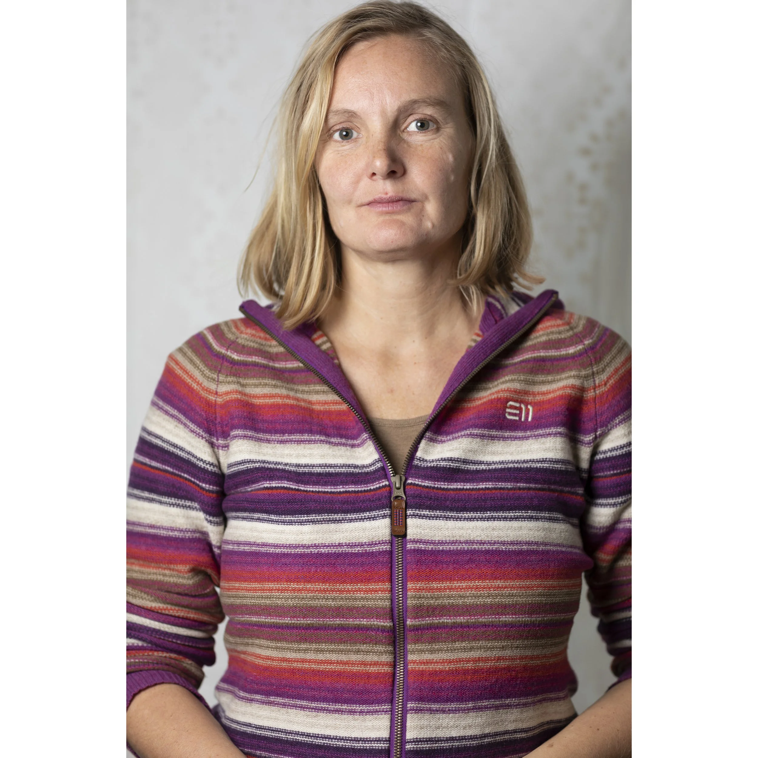 Porträt einer Frau mit blondem Haar, die einen gestreiften Pullover trägt, vor einem hellgrauen Hintergrund.