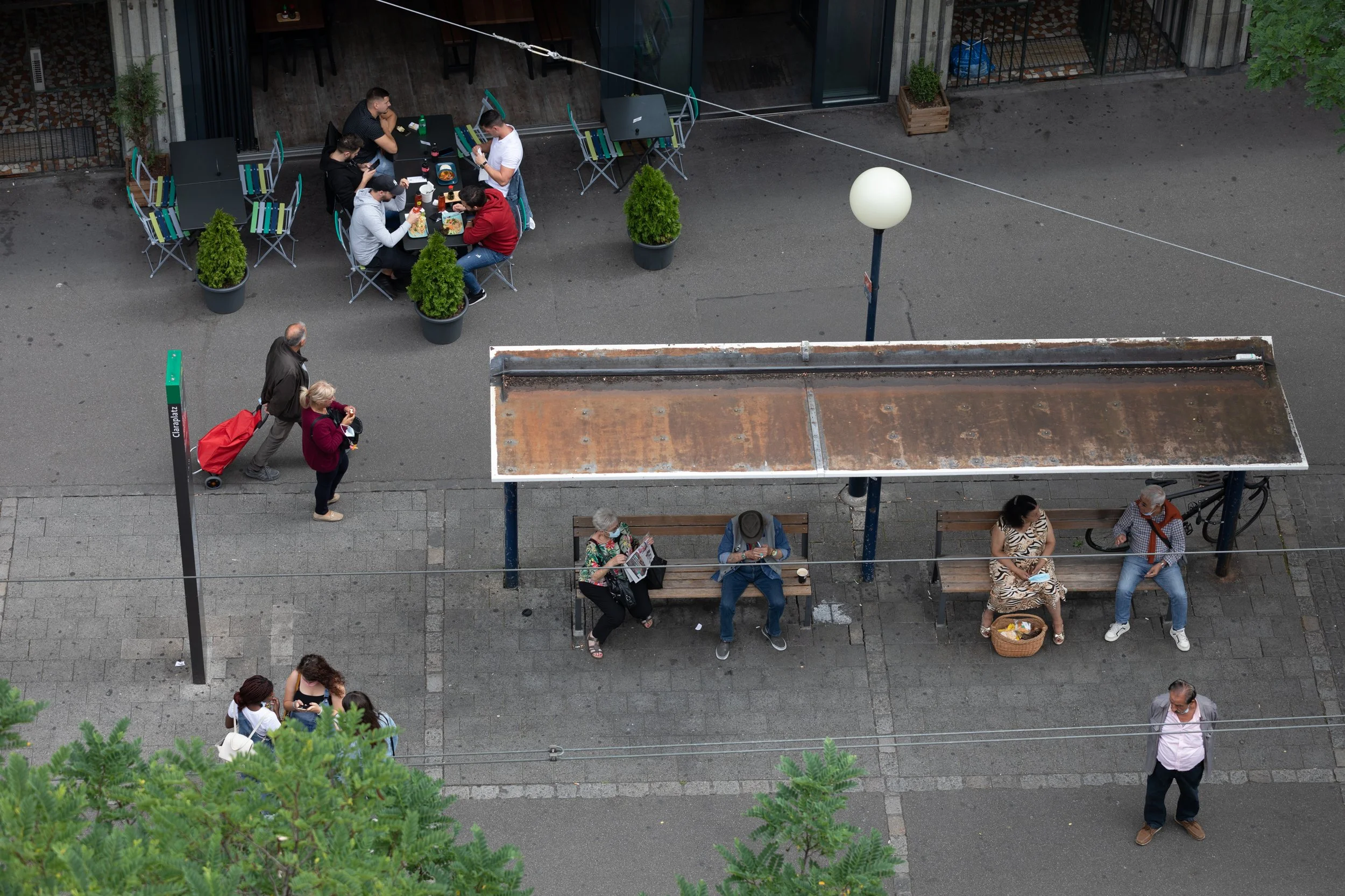 Aufnahme von oben auf eine Straßenszene mit mehreren Menschen, die auf Bänken sitzen, Leute, die vorbeigehen, und Tischen im Außenbereich eines Cafés oder Restaurants. Es gibt auch eine Bushaltestelle und einige Pflanzen.