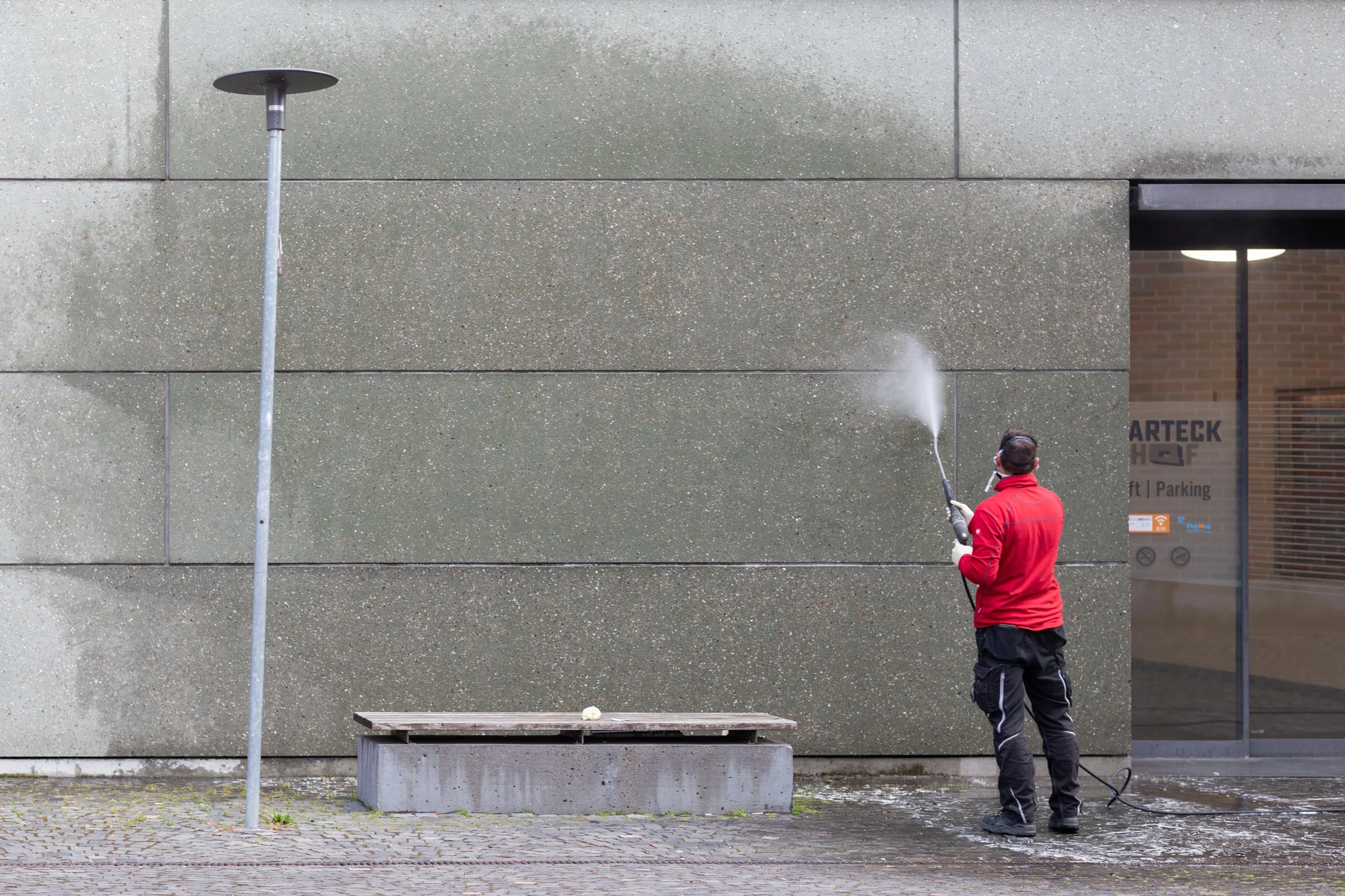 Ein Mann in roter Jacke säubert eine graue Betonmauer mit einem Hochdruckreiniger, darunter eine Parkbank und ein Aufsteller mit einem Licht.