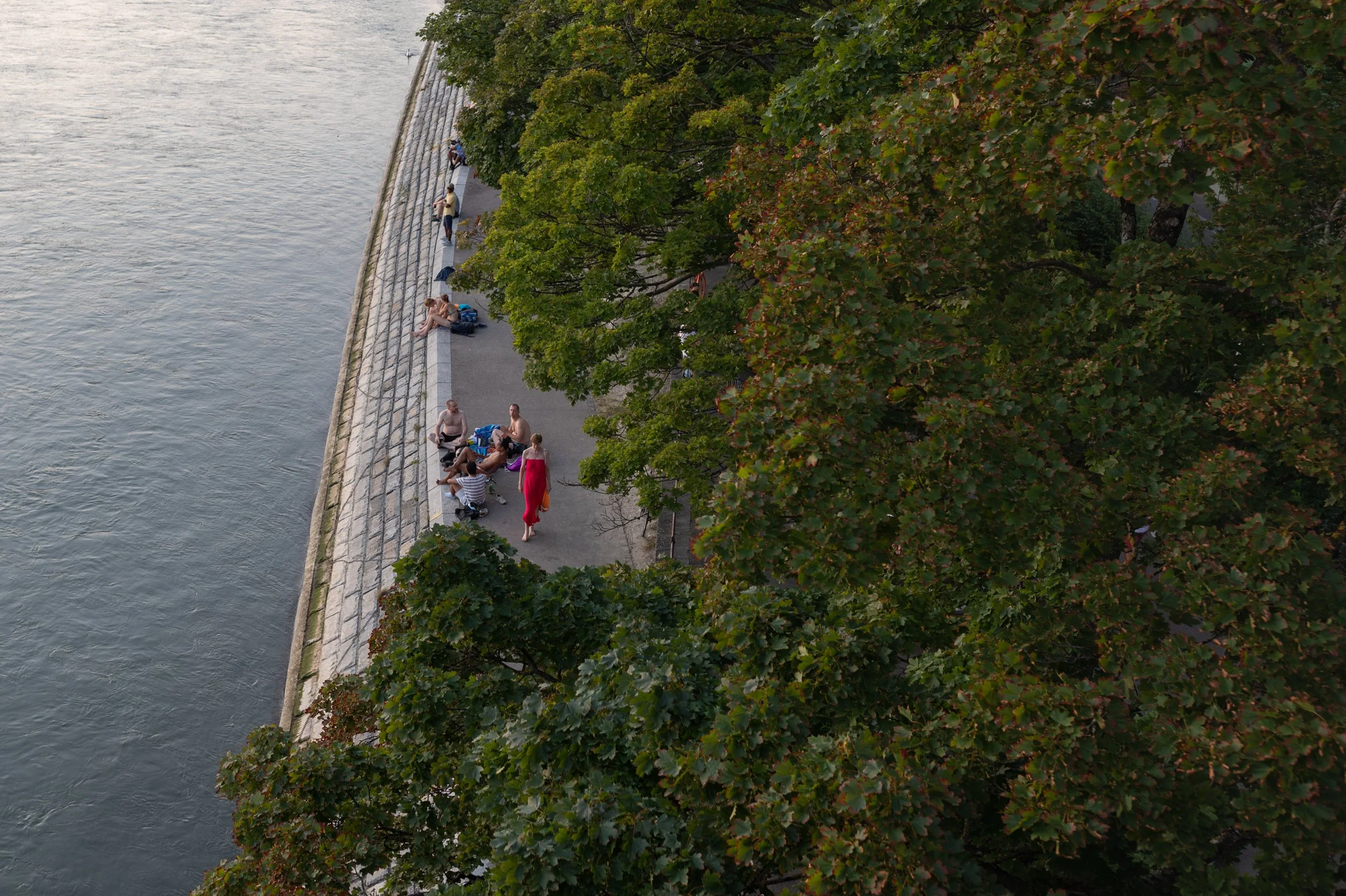 Blick auf einen Fluss mit einer Uferpromenade, auf der Menschen sitzen und spazieren gehen, sowie grüne Bäume, die die Szene umranden.