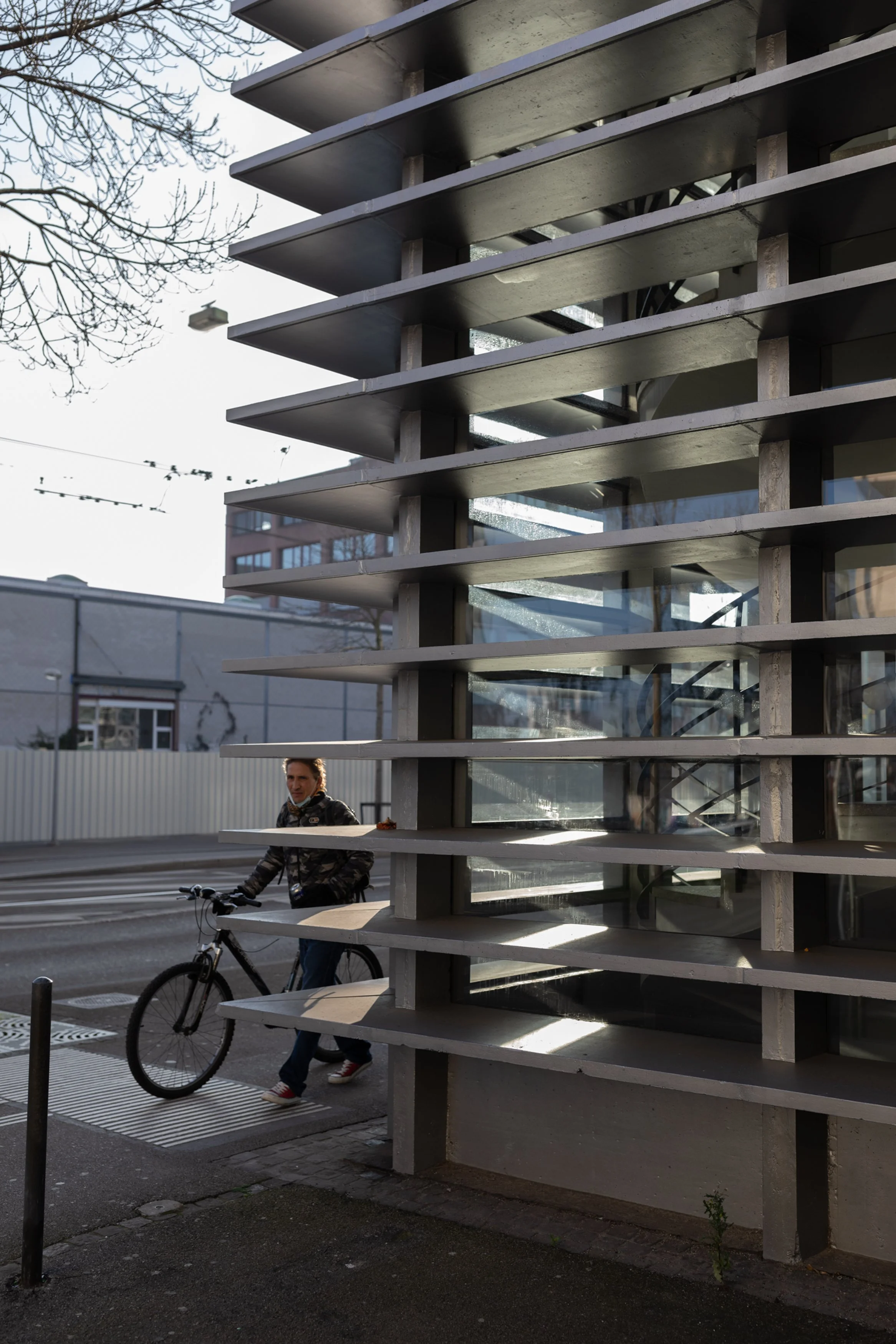Ein mehrstöckiges Gebäude mit horizontalen Balkonen und großen Glasfenstern. Im Vordergrund ein Mann auf einem Fahrrad, der an einer Frau vorbeifährt, die neben dem Fahrrad auf dem Boden steht.