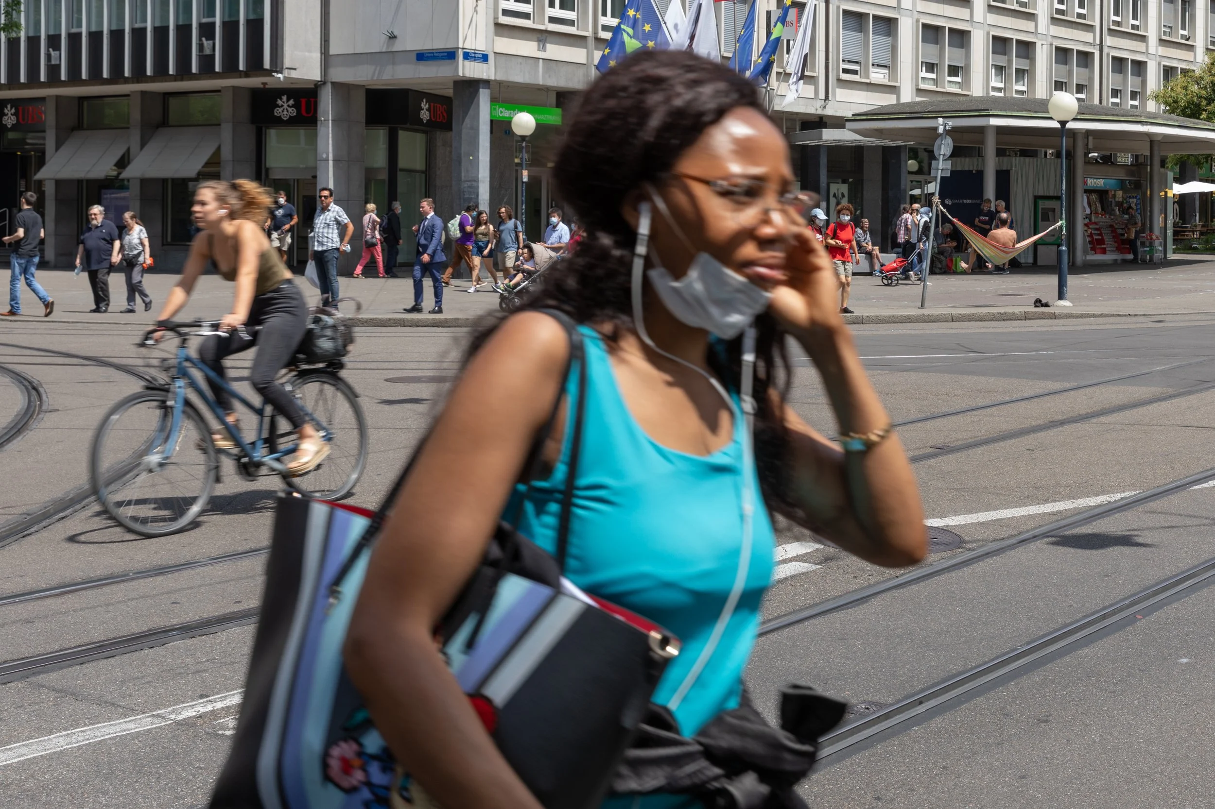 Straßenansicht mit Menschen, die Fahrrad fahren und zu Fuß unterwegs sind, im Hintergrund ein Bürogebäude mit Flaggen und Geschäften wie UBS und Clearstream.