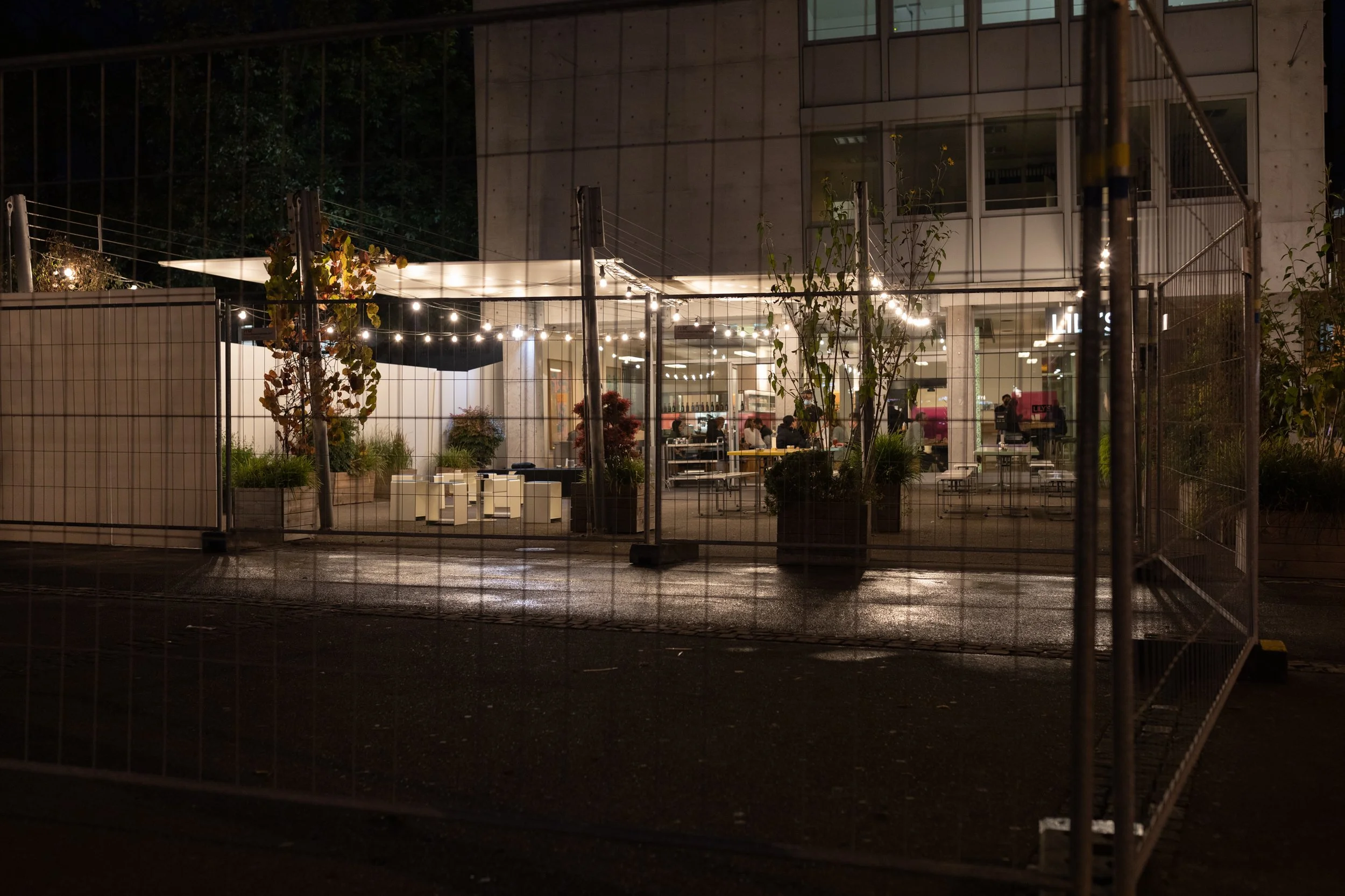 Ein beleuchteter gastronomischer Bereich hinter einem Metallzaun bei Nacht, mit Tischen und Menschen inside.