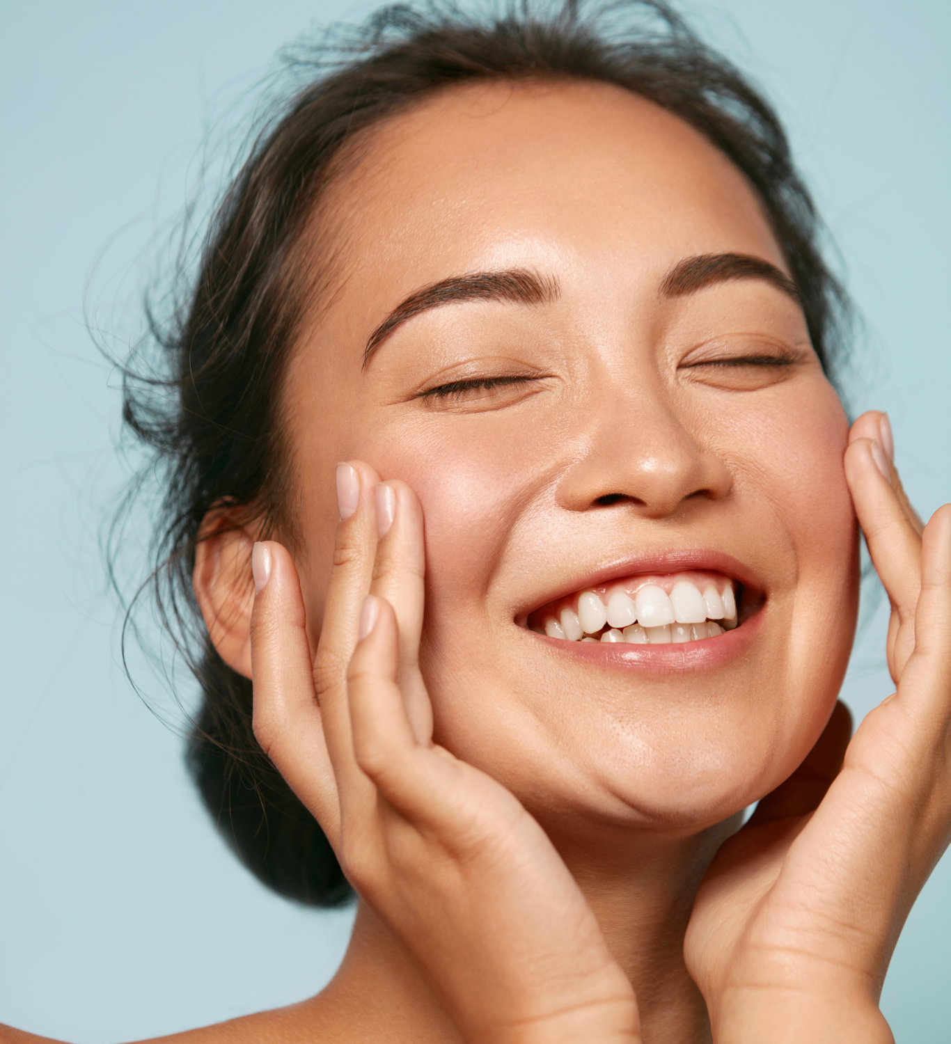 A woman with dark hair smiling and touching her face gently, against a light blue background.
